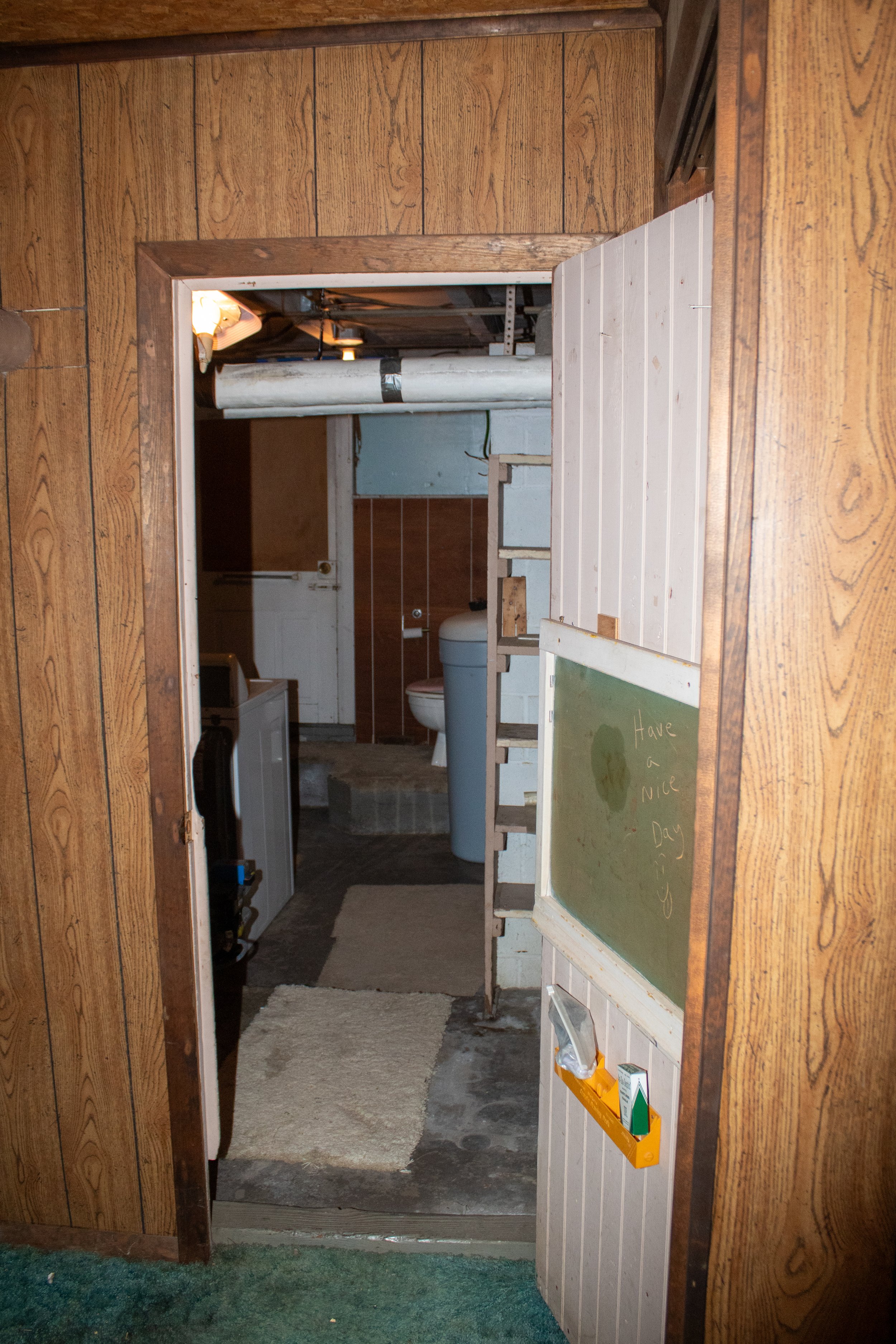 Old basement with wood paneling, a doorway leading to a utility area with exposed pipes, water heater, and a toilet. A chalkboard with 'Have a nice day' written on it is visible.