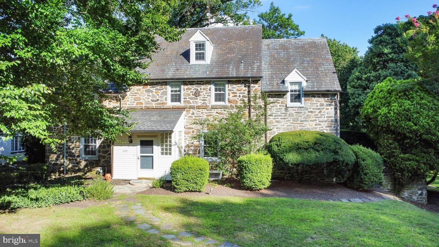 Stone house with gabled roof, surrounded by trees and shrubs in a grassy yard.