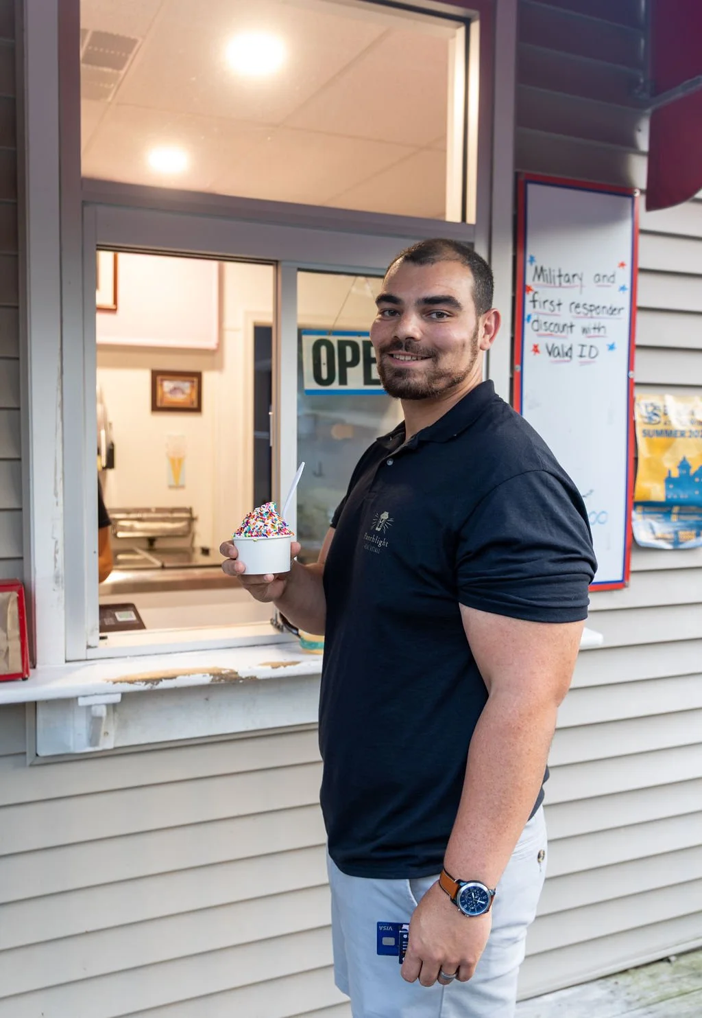 A man with a beard and short hair wearing a navy polo shirt and white shorts, holding a bowl of ice cream with colorful sprinkles, standing outside an ice cream or snack shop with a window and an 'OPEN' sign.