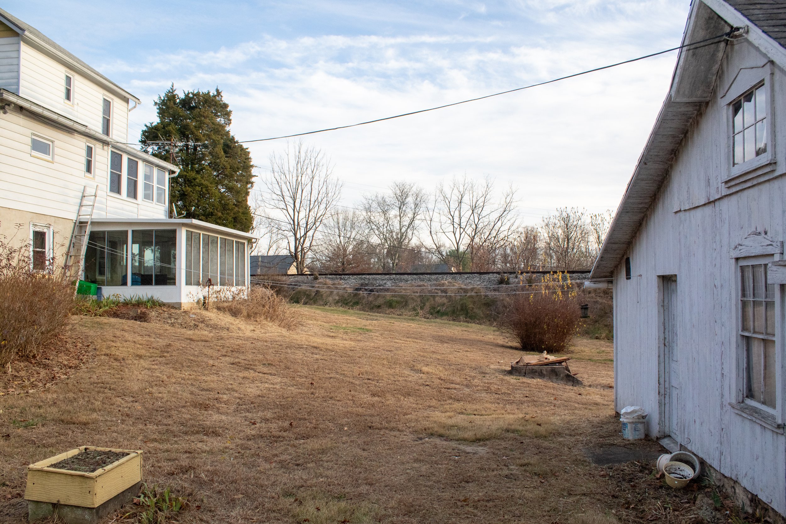 Backyard with a white house, connected sunroom, and a detached white building, surrounded by dry grass and bare trees.