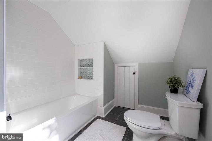 Modern bathroom with white bathtub, gray walls, small window, toilet, and decorative plant on tank lid. Slate tile floor and white bath mat accentuate minimalist design.