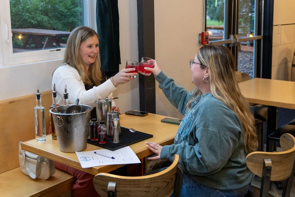 Two women cheers their cocktails they made at the Porchlight Real Estate Team's Cocktail workshop. they're sitting at a table together where they found something to do on the weekend and have a pen and paper to win trivia