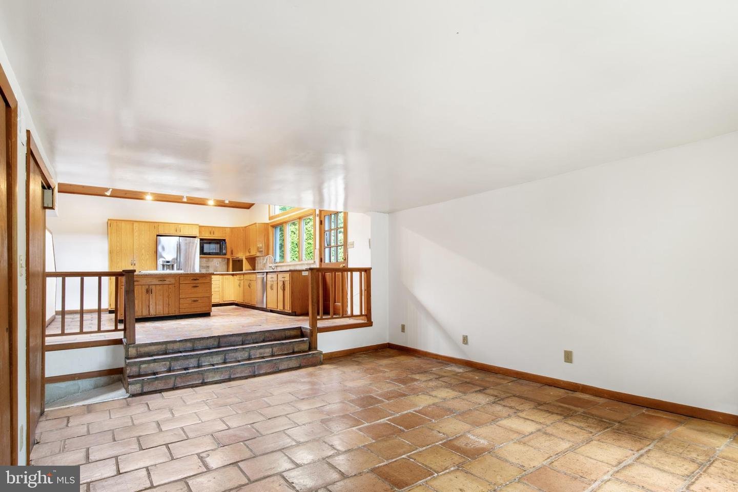 Spacious room with terracotta tile flooring leading to a kitchen area with wooden cabinets, a stainless steel refrigerator, and a large window. The kitchen is elevated with a few steps leading up to it and features a wooden railing.