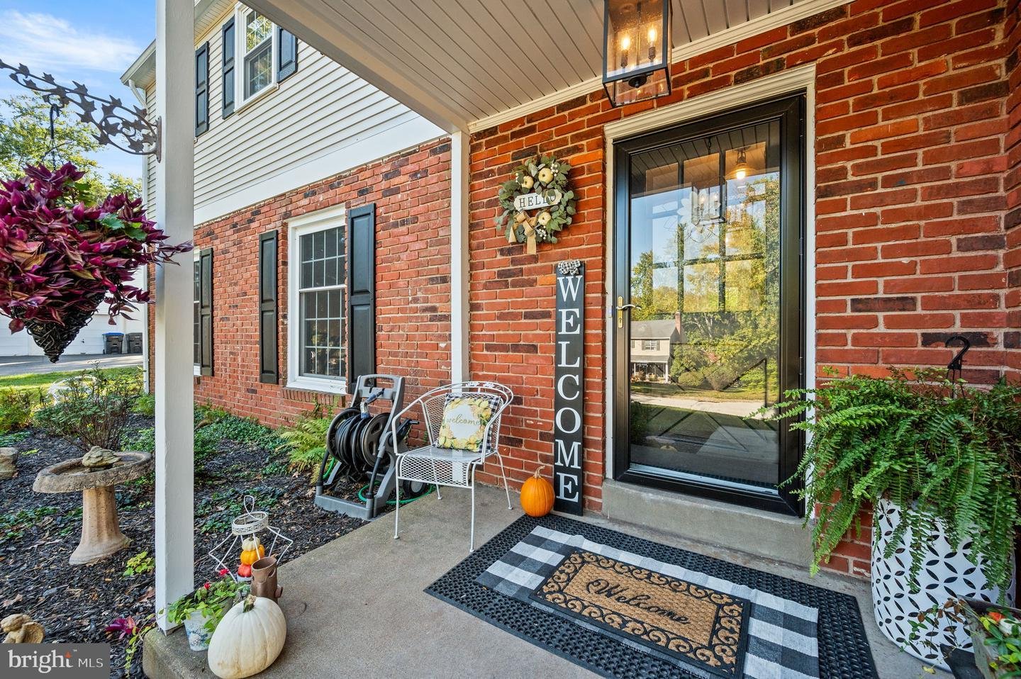 Front porch of a brick house with a black door, welcome sign, mat, and seasonal decorations including pumpkins and hanging plants.