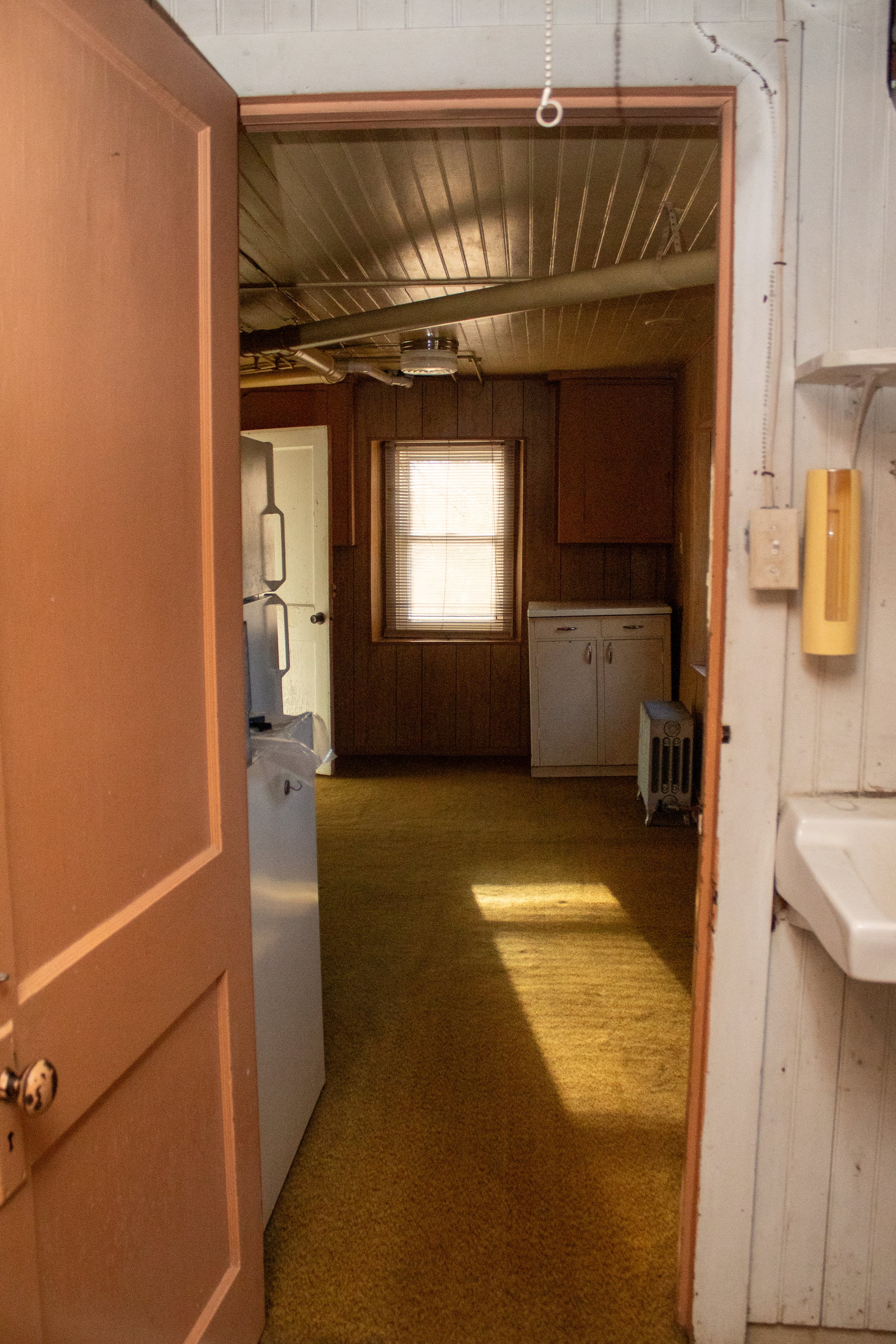 Interior view of a vintage kitchen with wood paneling, a window with blinds, an open wooden door, and faded yellow carpet. A refrigerator and sink are visible.