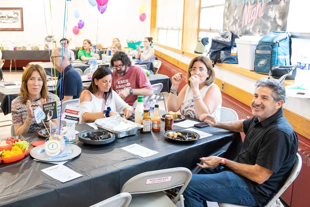 Guest sit at decorated tables enjoying food and conversation during the Porchlight Real Estate Team's trivia night fundraiser for Maccabi youth sports, with colorful balloons and trivia signs adding to the lively atmosphere
