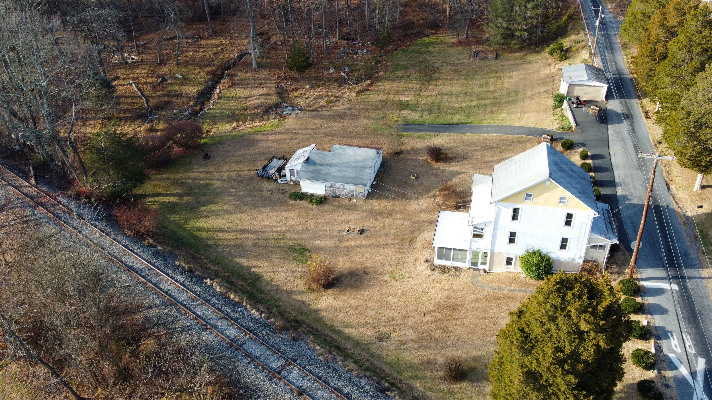 Aerial view of a rural area featuring a white house with a yellow accent, adjacent outbuildings, and a driveway leading to a paved road. There is an open yard with some trees and a nearby railroad track. Surrounding the property is a wooded area with