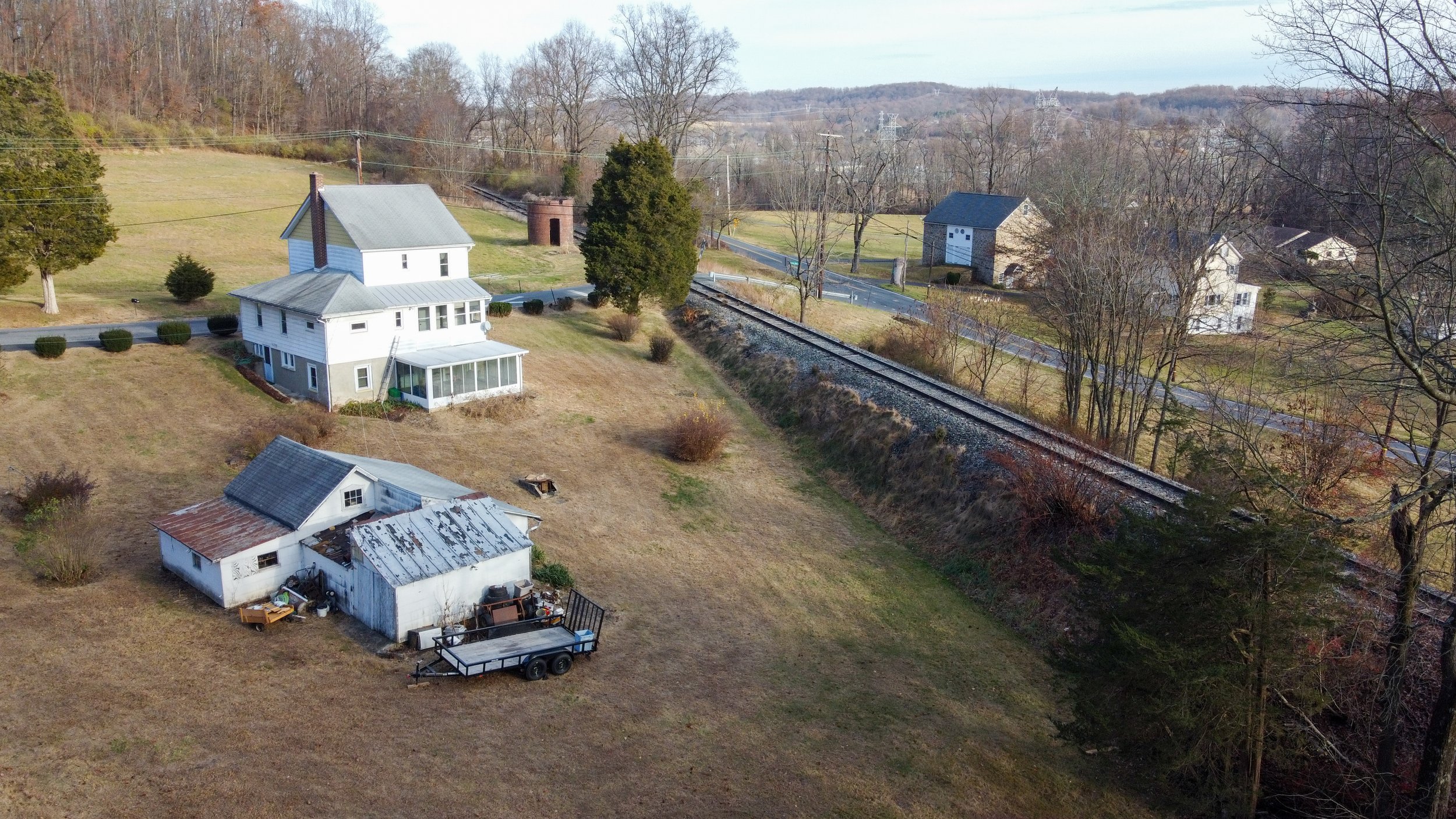 Aerial view of a rural landscape with a white house, a small outbuilding, and a trailer. A railway track runs parallel to a country road. The area is surrounded by fields, trees, and distant hills.