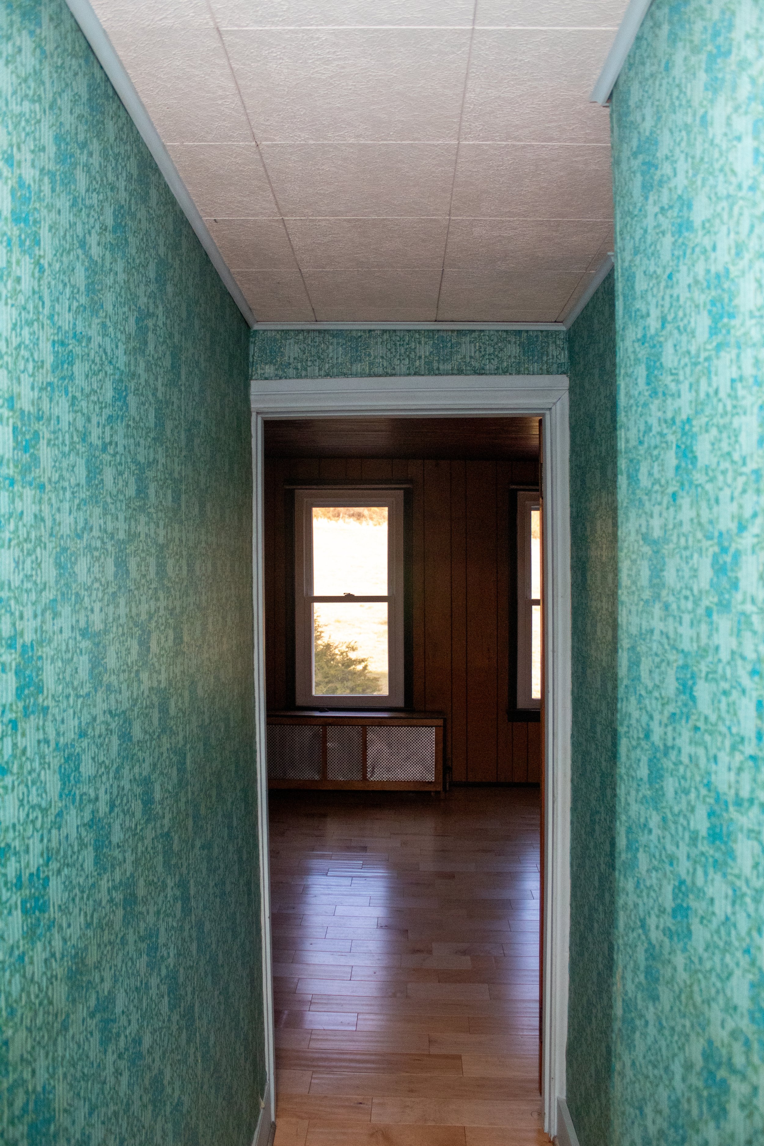 A narrow hallway with green patterned wallpaper leading to a room with wood-paneled walls, two windows, and a wooden floor.