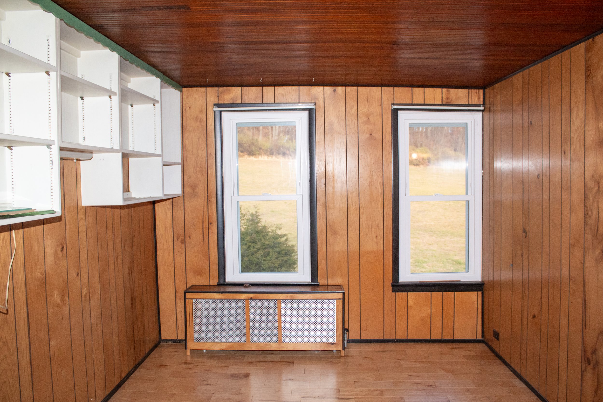 Empty room with wood paneling, two windows, white shelving, and a radiator cover under the windows.