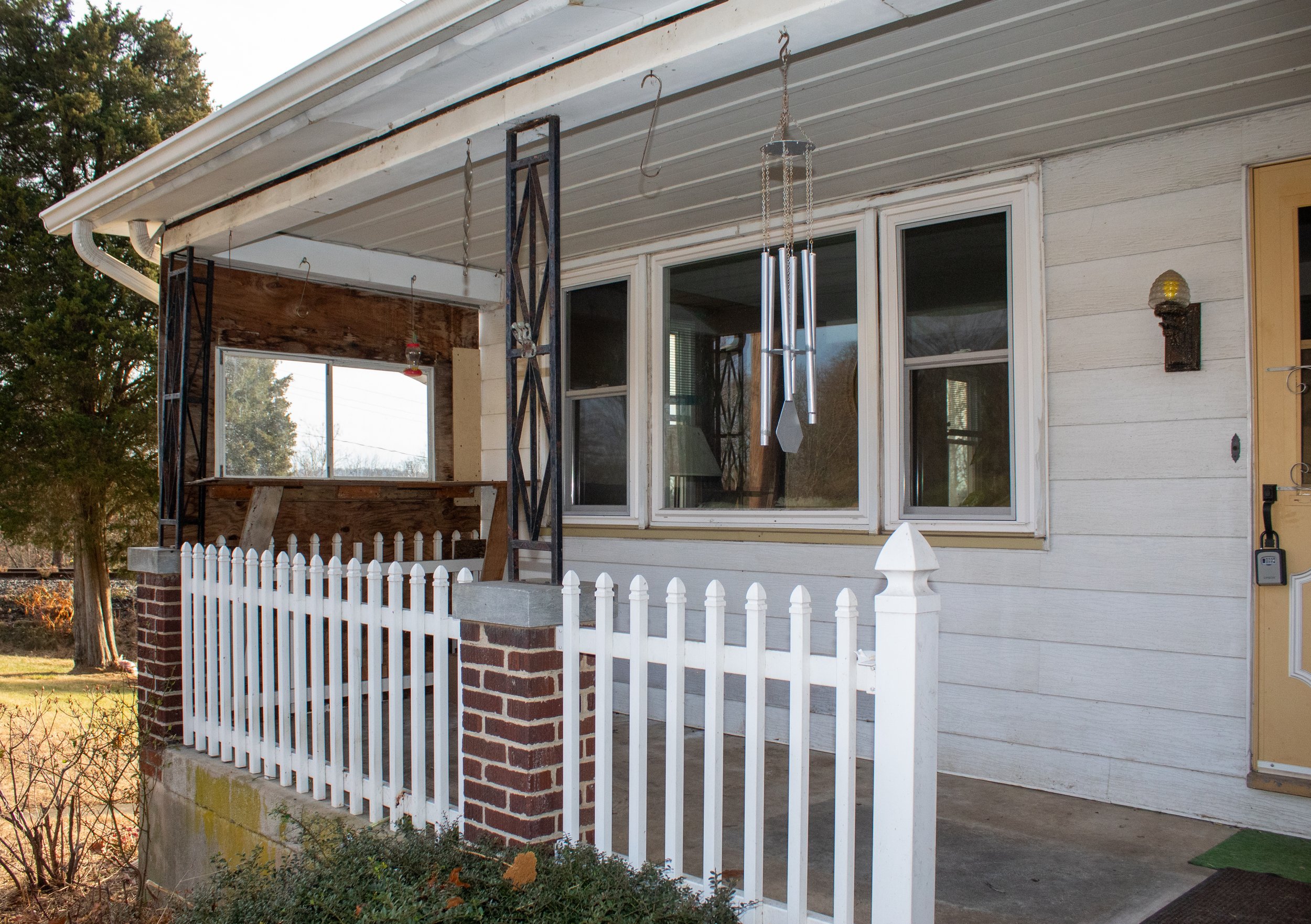 Front porch of a house with a white picket fence, metal hanging hooks, wind chime, and a yellow door.
