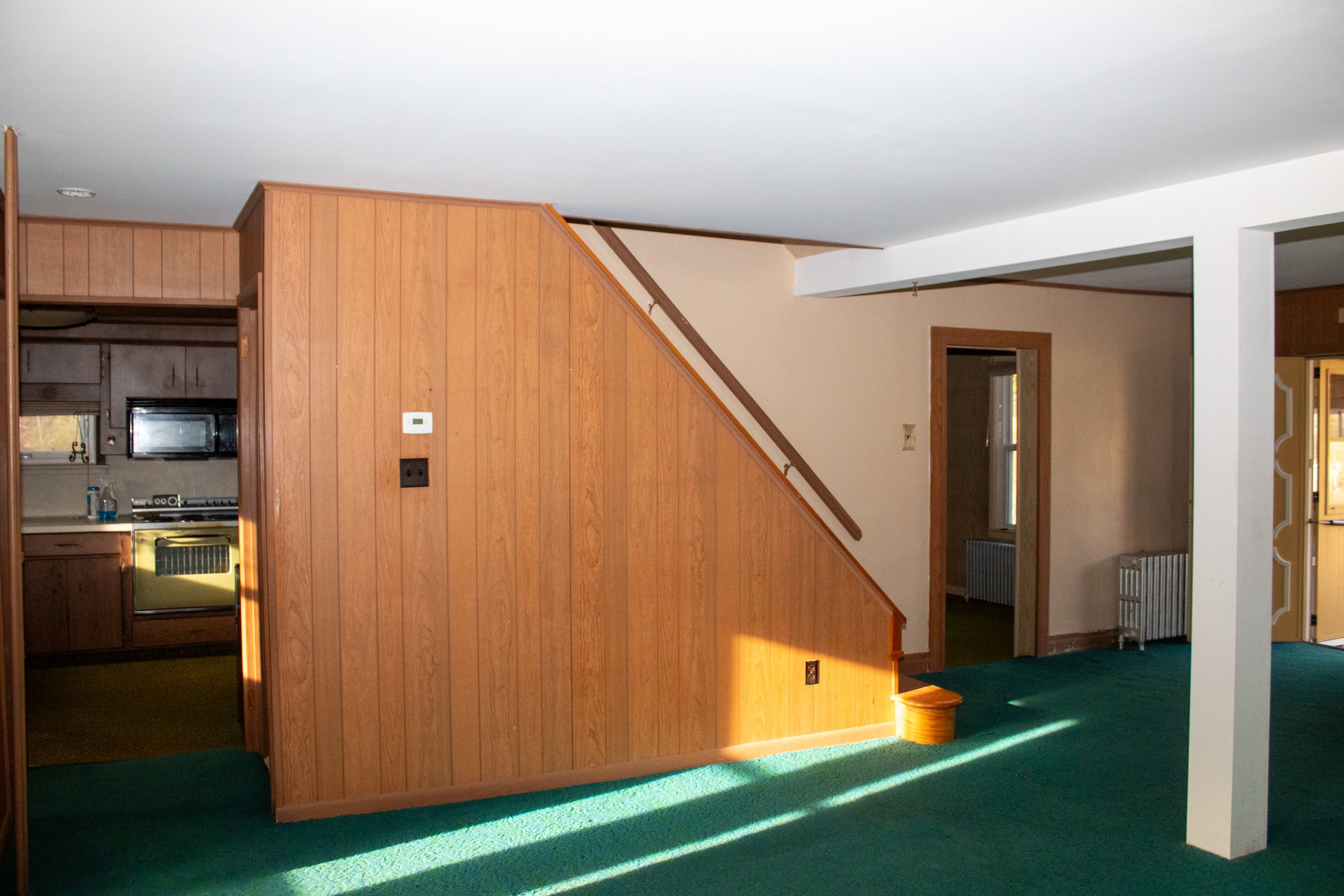 Interior of a room with wooden wall panels, green carpet, and a visible staircase. A kitchen area with a stove and microwave is partially seen on the left. There's a doorway leading to another room on the right.