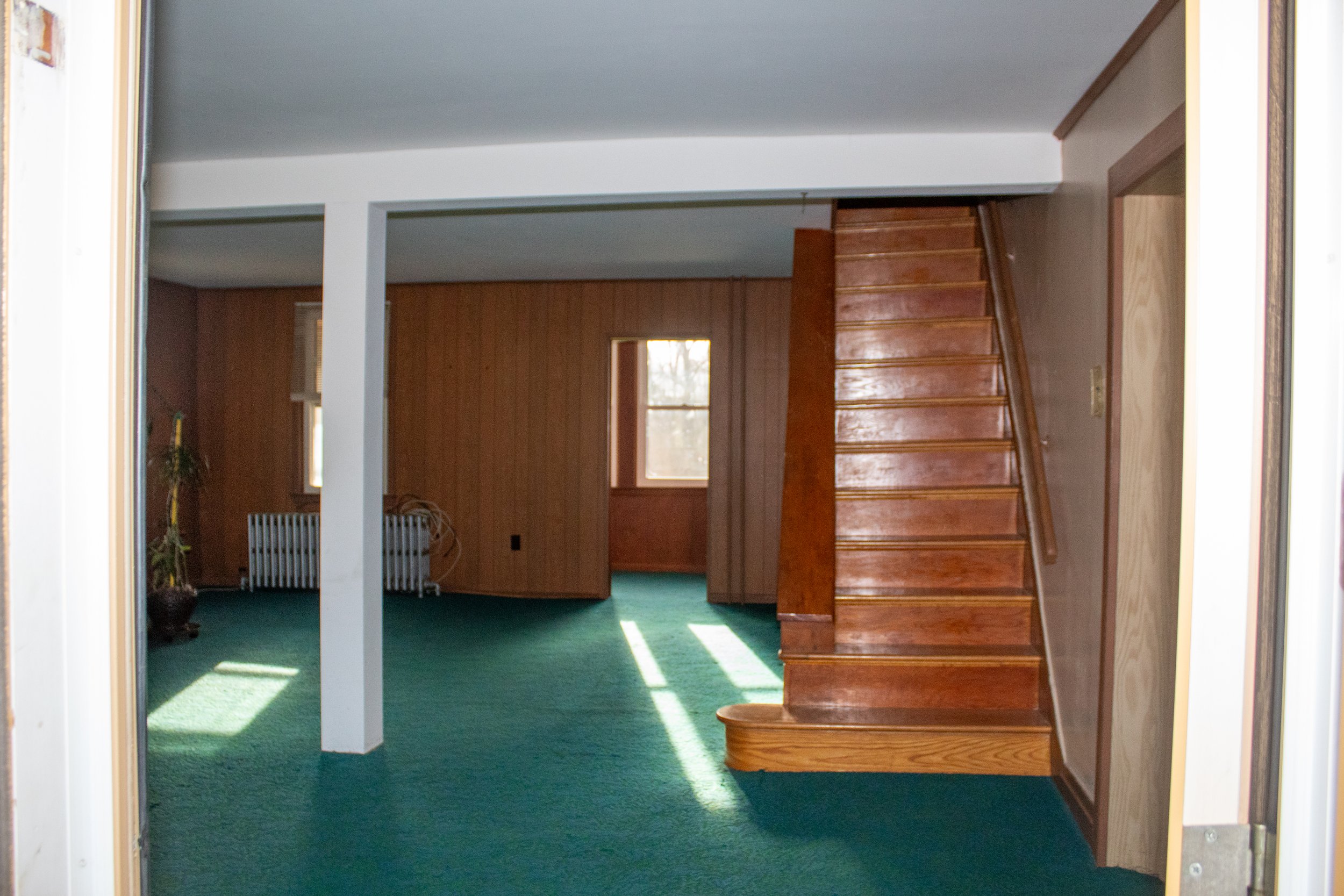 Interior room with green carpet, wooden staircase, radiator, and windows; wood-paneled walls.