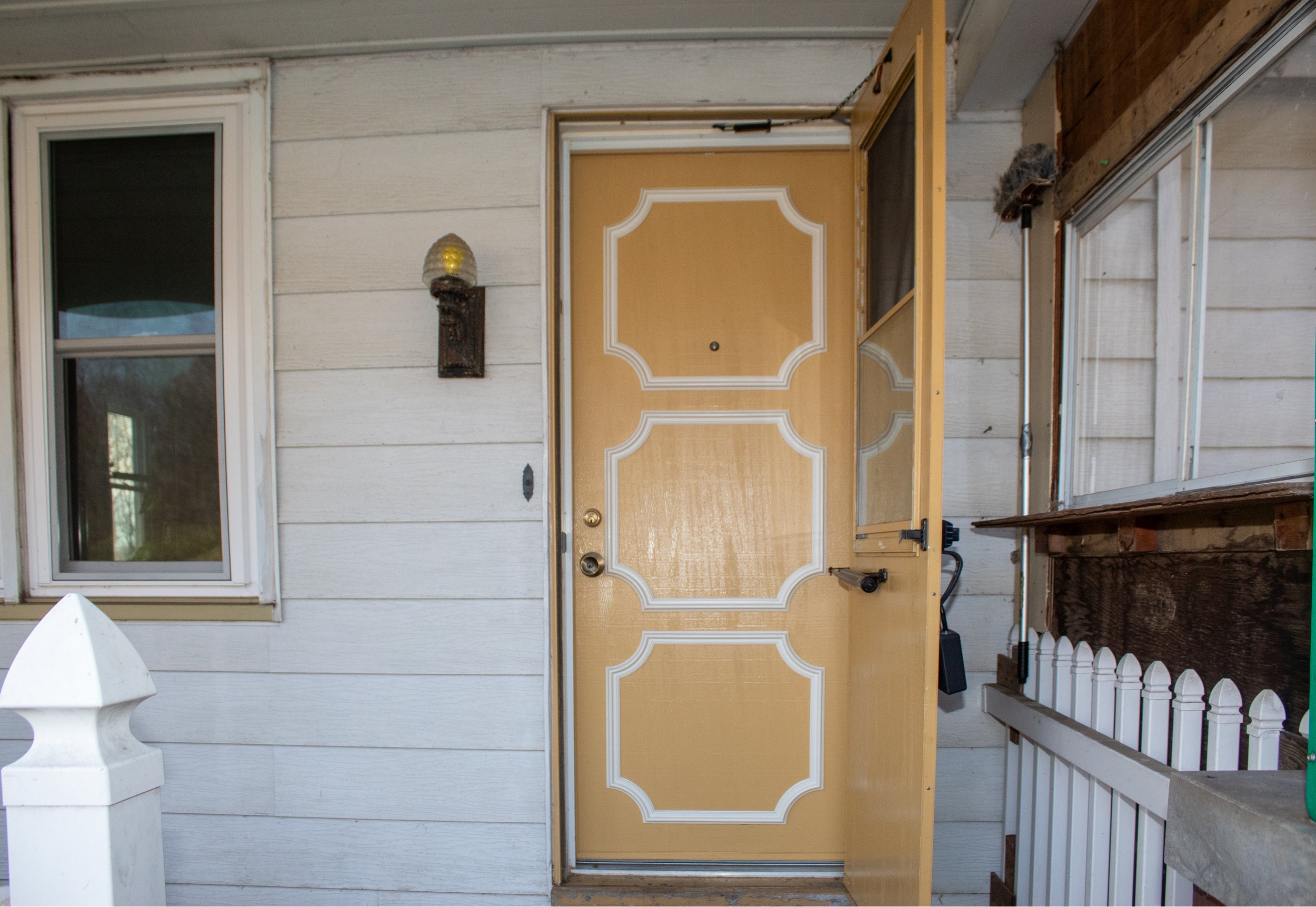 Front porch with a beige wooden door, white detailing, a black handle, a glass storm door, and a nearby lantern. Adjacent is a window and white wooden paneling.