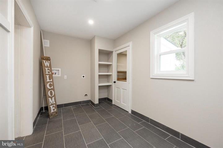 Mudroom interior with gray tiled floor, beige walls, small window, open shelves, and a "Welcome" sign leaning against the wall.
