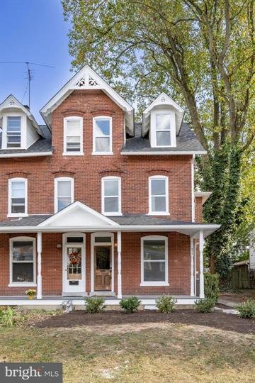 Three-story brick house with white trim, a front porch, and a gabled roof, surrounded by greenery and trees.