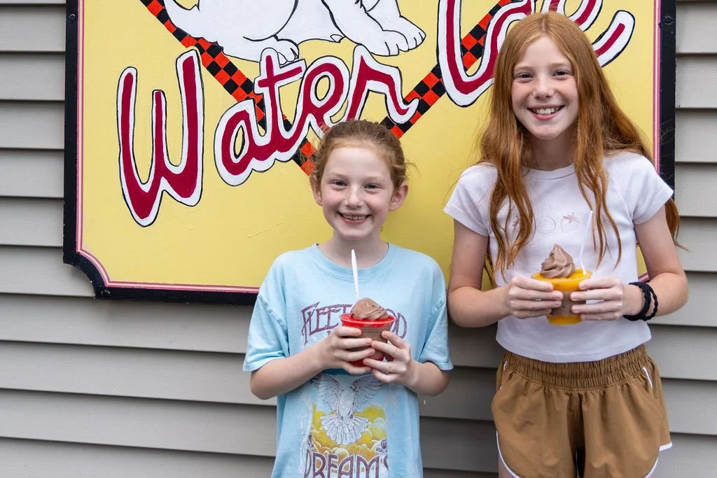 Two smiling girls stand in front of Annie's Water Ice Sign in DOylestown Holding cups of chocolate soft serve and water ice during a Porchlight Real Estate Team Community Event offering free water ice to raise money for local community programs