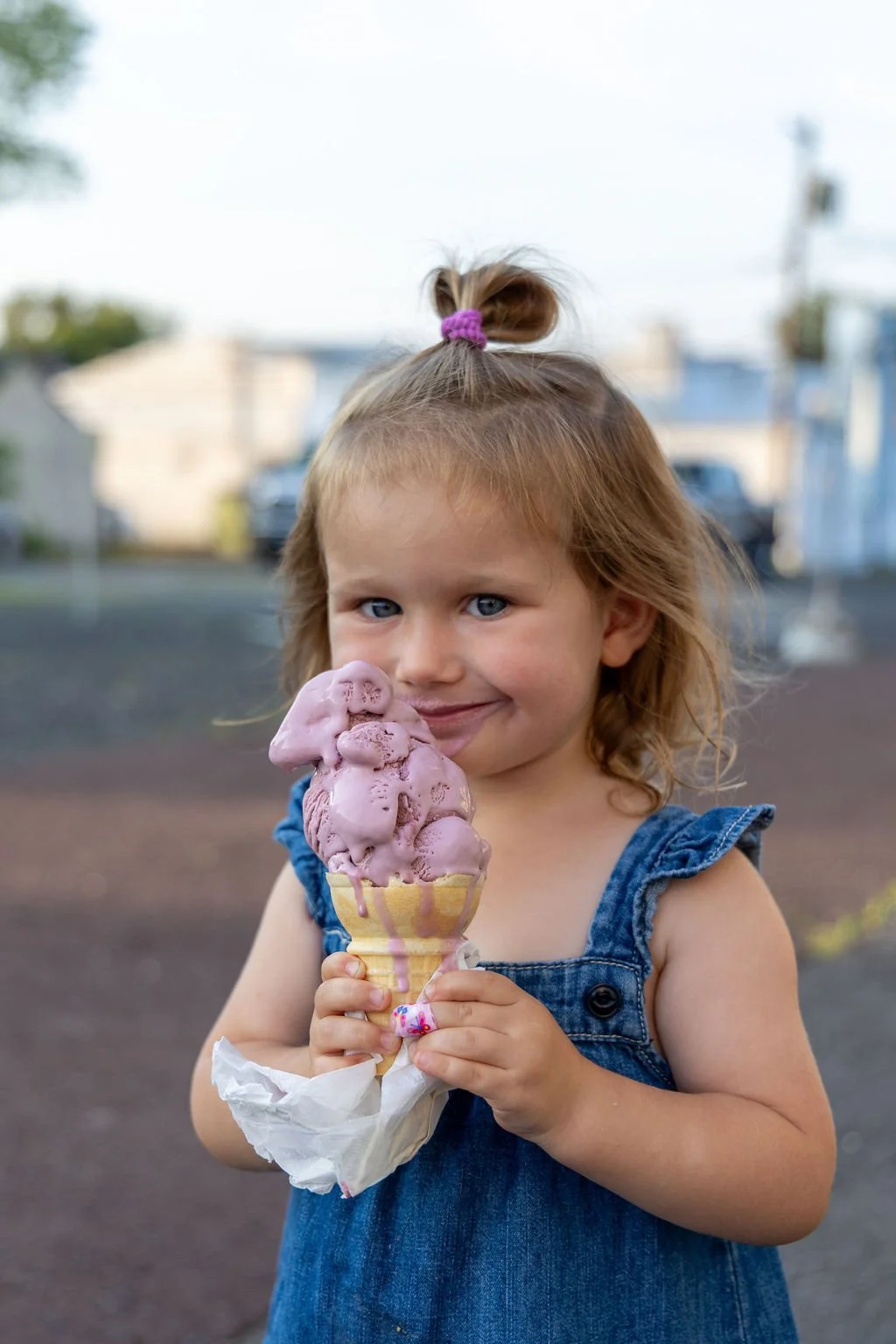 A young girl with light brown hair tied in a top knot, holding a large ice cream cone with pink ice cream, standing outdoors with a blurred background of a street and buildings.
