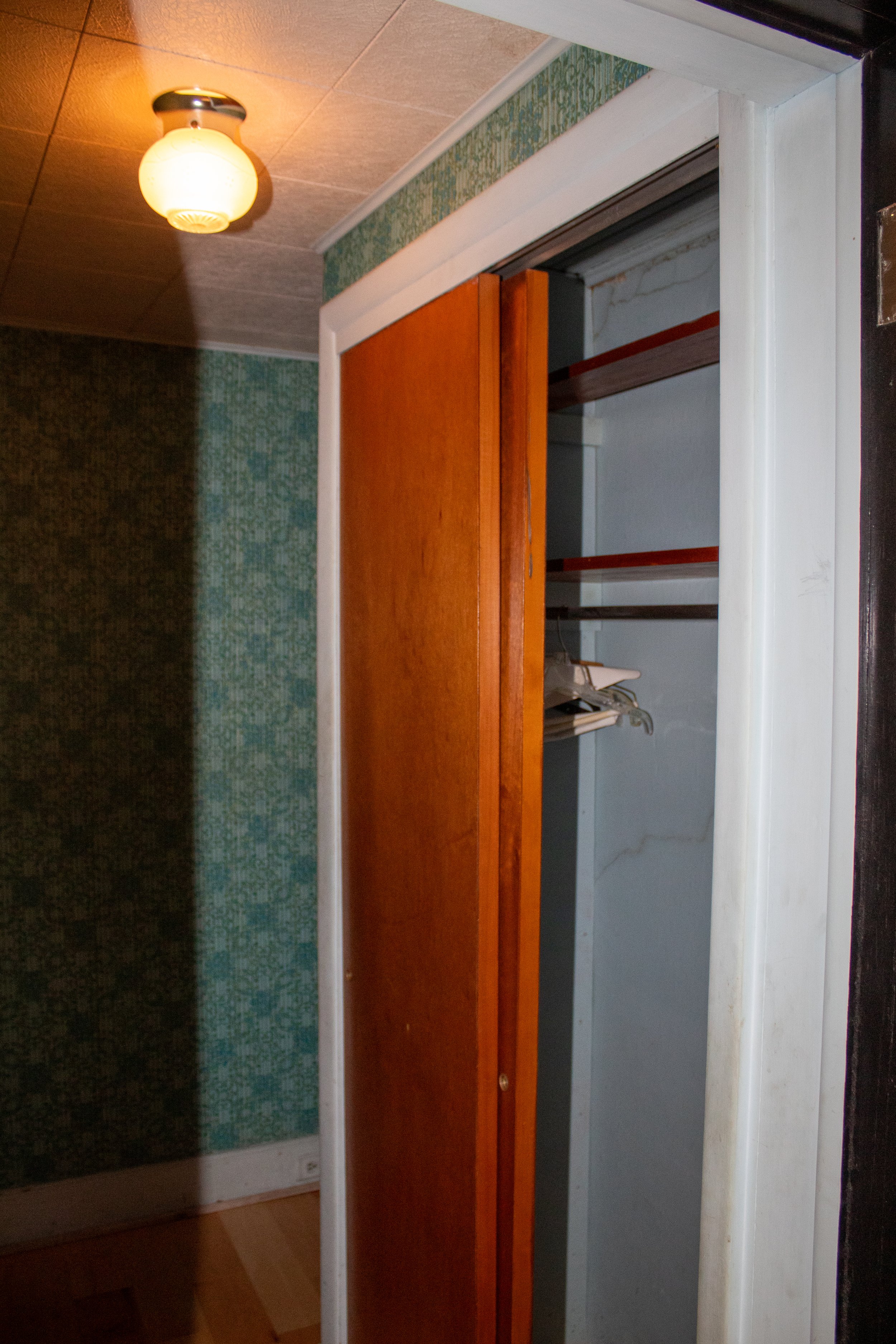 Vintage closet with wooden sliding door, empty hangers, and overhead light fixture in a room with patterned wallpaper.