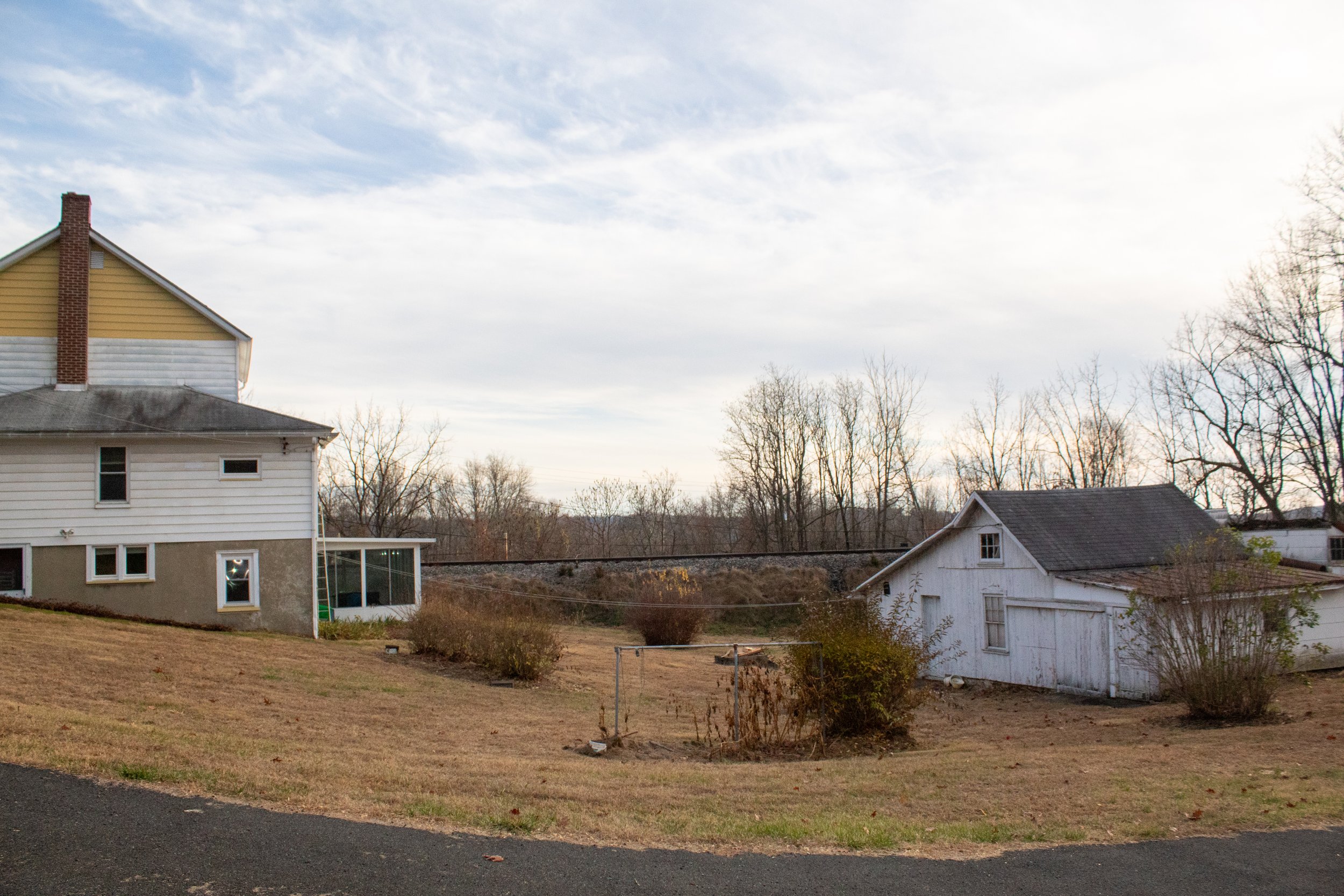 A rural landscape featuring a large house with a chimney and a smaller white building, possibly a barn or shed, on a grassy area with some bushes. The sky is cloudy, and leafless trees are in the background.