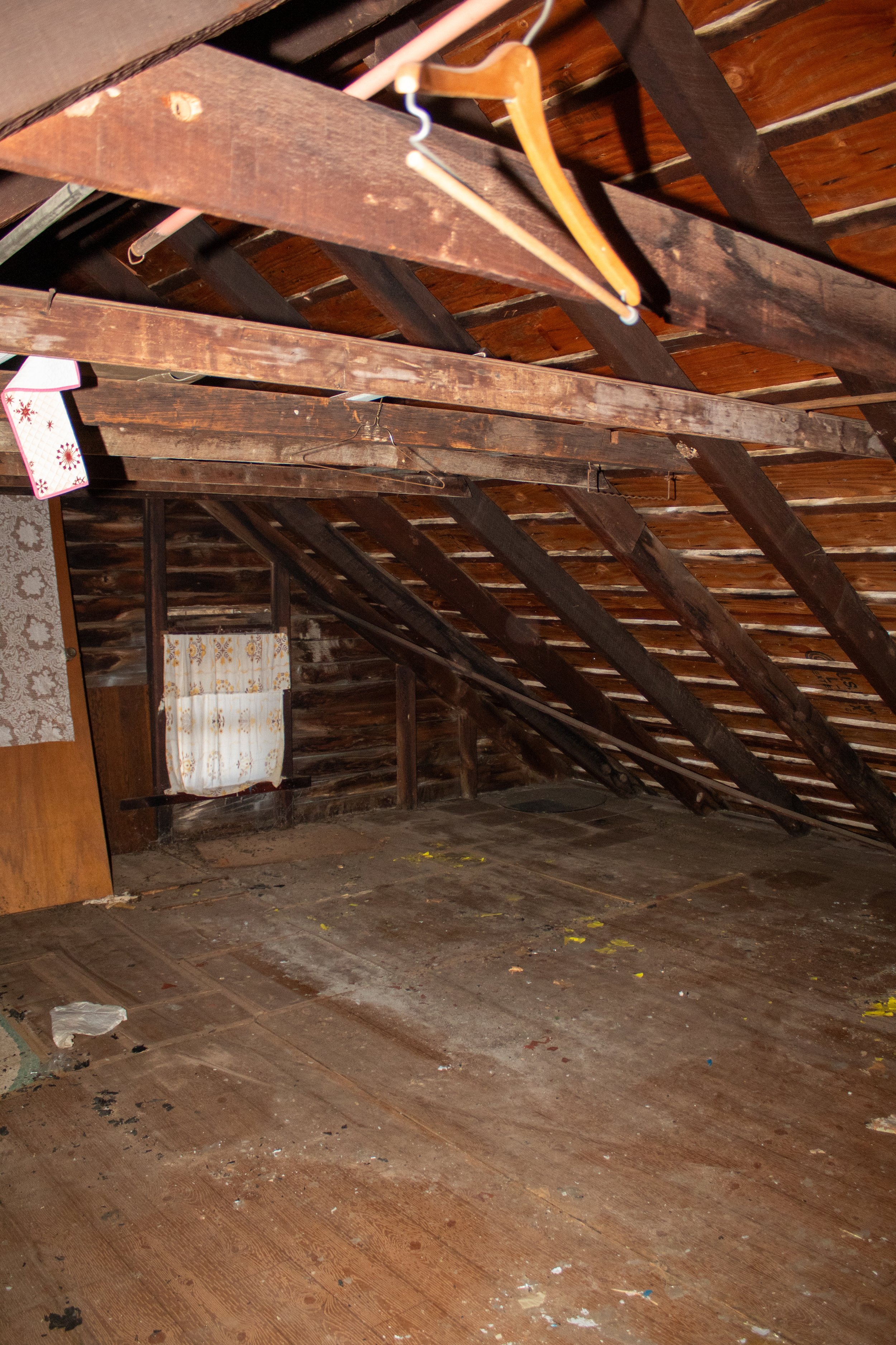 Old dusty attic with exposed wooden beams, hangers, and a small curtain-covered window.