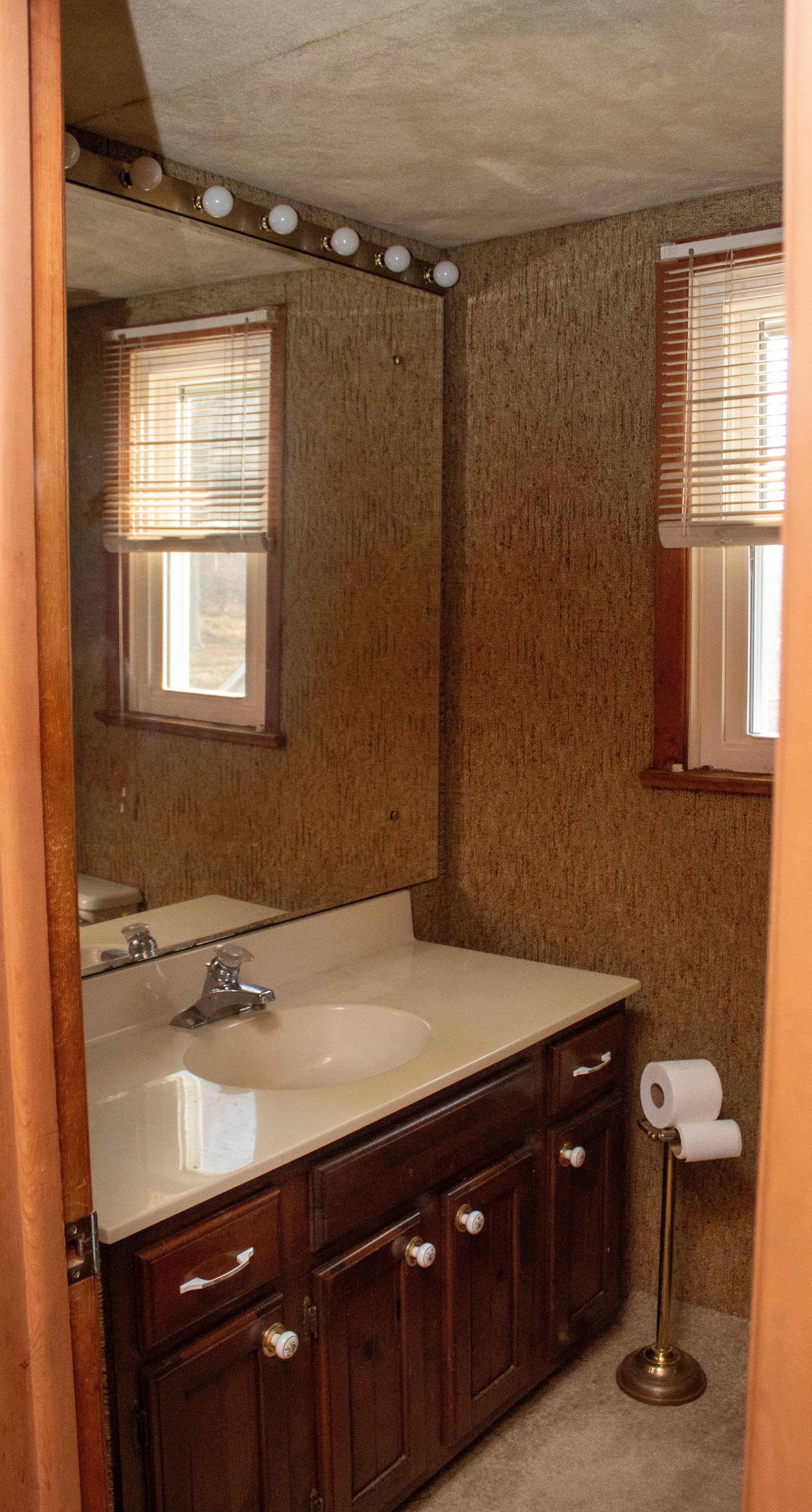 Bathroom with wooden cabinet, countertop sink, mirror, and window with blinds. Light bulbs line the top of the mirror, and a toilet paper holder stands nearby.