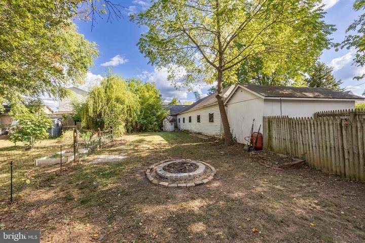 Backyard with a brick fire pit, grassy area, trees, white fence, and a white building in the background under a blue sky.