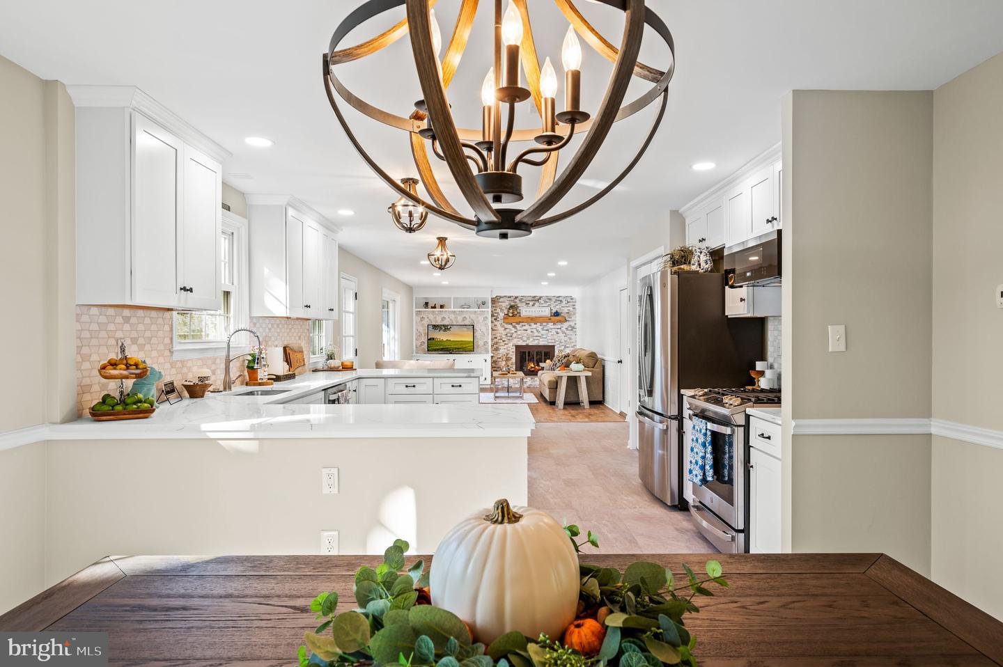 Modern kitchen and living area with white cabinets, stainless steel appliances, overhead lighting fixture, and fall-themed table centerpiece.