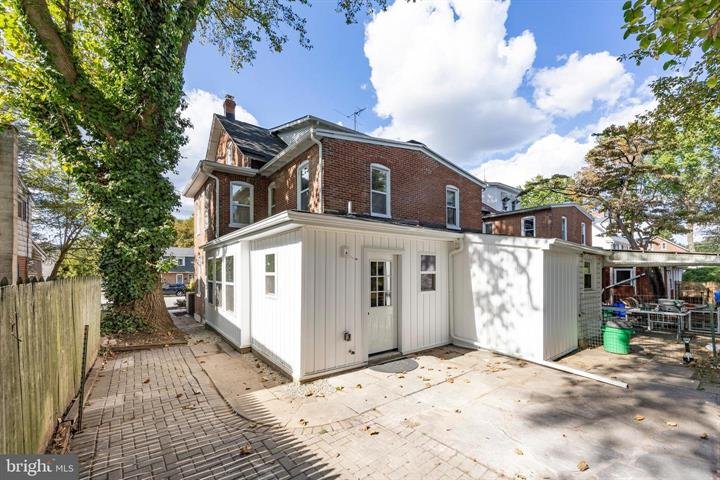 Brick house with white extension and paved backyard, surrounded by trees, under a partly cloudy sky.