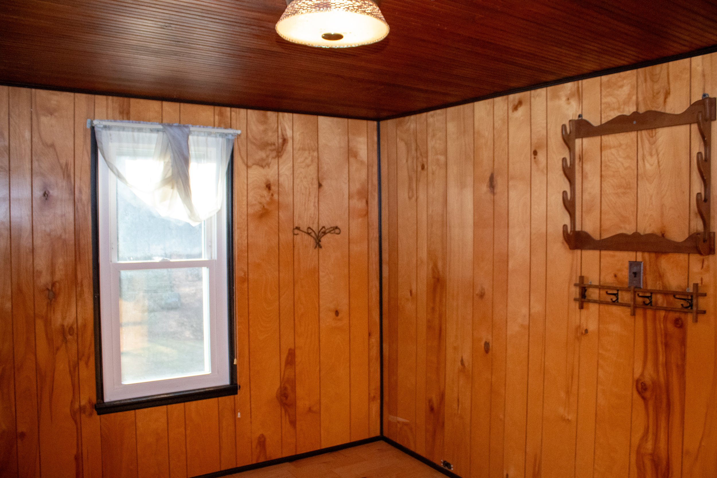 Wood-paneled room with a window, ceiling light, curtain, and decorative wall hooks.