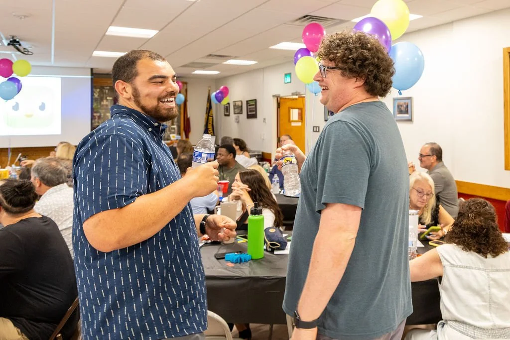 two men laughing and talking while holding water bottle during the Porchlight Real Estate Team's Trivia Night fundraiser for Maccabi youth sports, surrounded by guests seated at tables decorated with colorful balloons