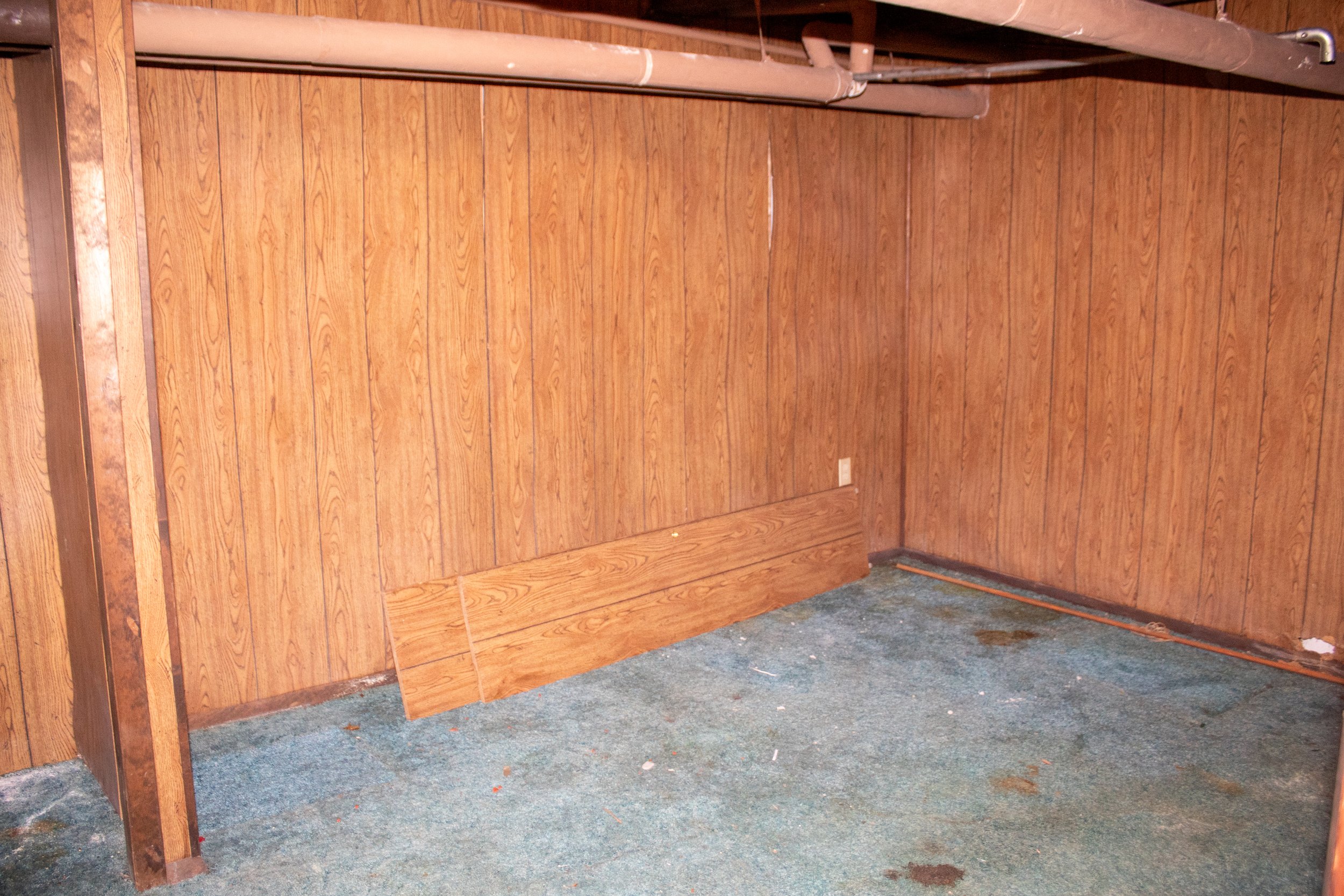 Empty basement room with wood-paneled walls, blue carpet, and exposed pipes on the ceiling.