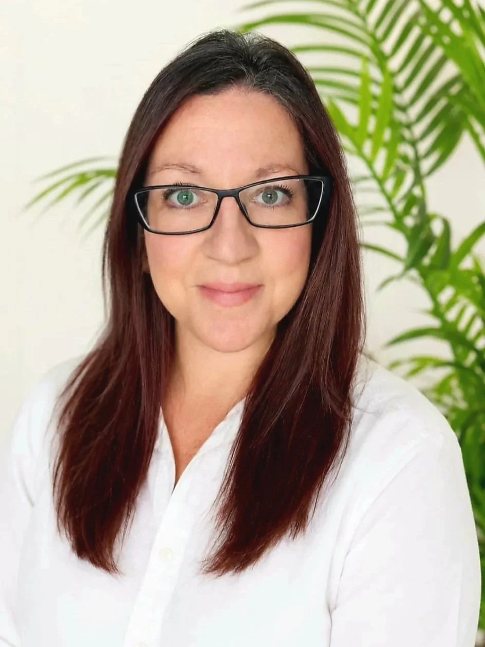 Rebekah, a Social Worker in Ontario, Canada serving Burlington, Ontario in person and various provinces. A woman with long brown hair, glasses, and fair skin, wearing a white shirt, smiling at the camera with a green leafy plant in the background.