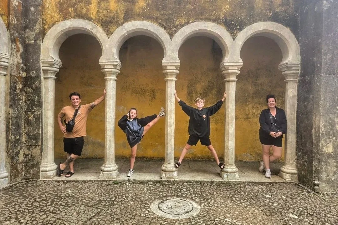 Four people posing inside a decorative stone architectural structure with three arches, on a cobblestone ground, against a weathered yellow wall.