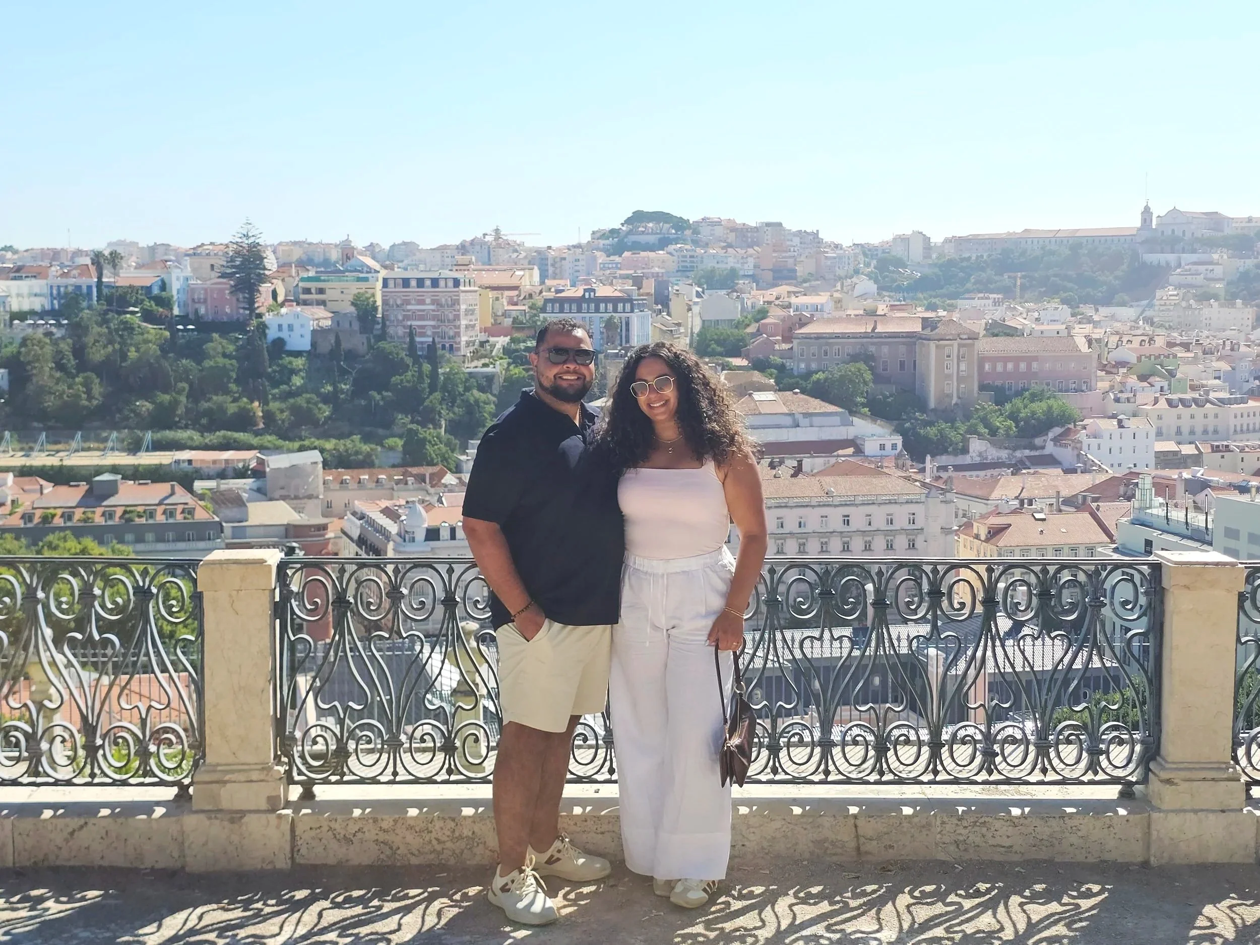 A smiling couple standing on a balcony overlooking a cityscape with hills, colorful buildings, and trees in the background.