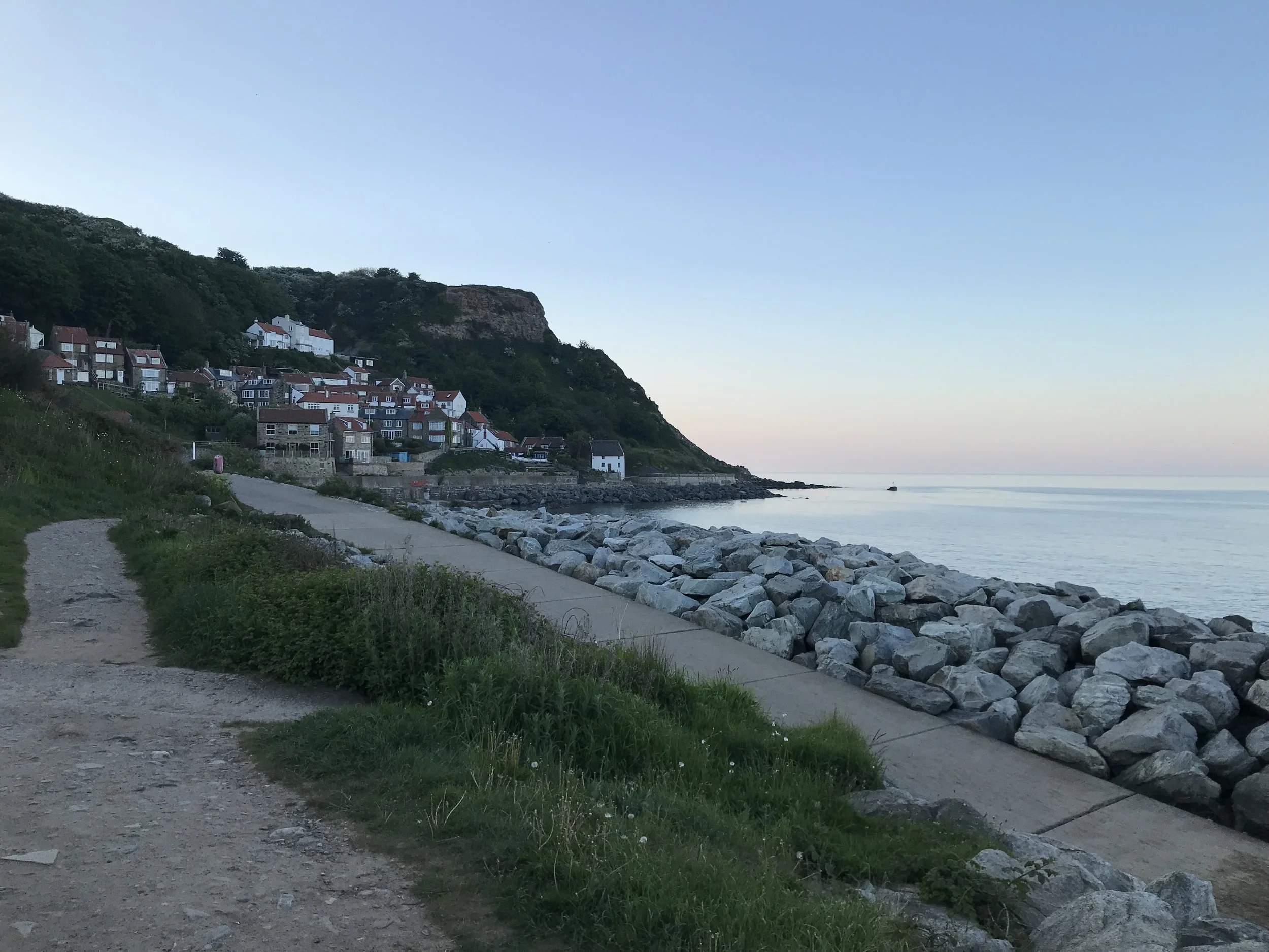 Seaside town with rocks along a walkway, houses on a hillside, calm water, and clear sky at sunset.