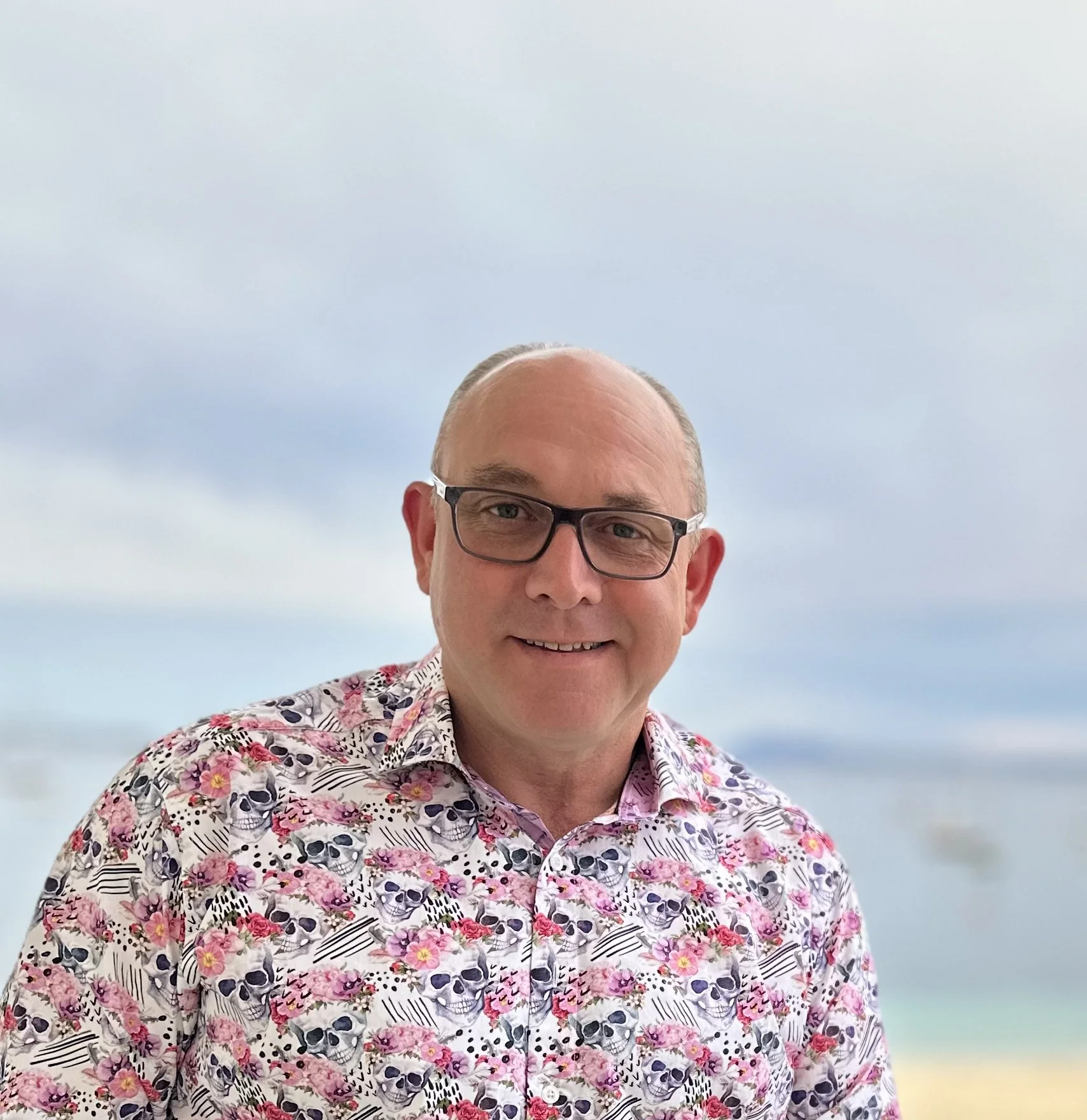 A man wearing glasses and a floral shirt with skull and flower pattern, smiling on a beach with the ocean and cloudy sky in the background.