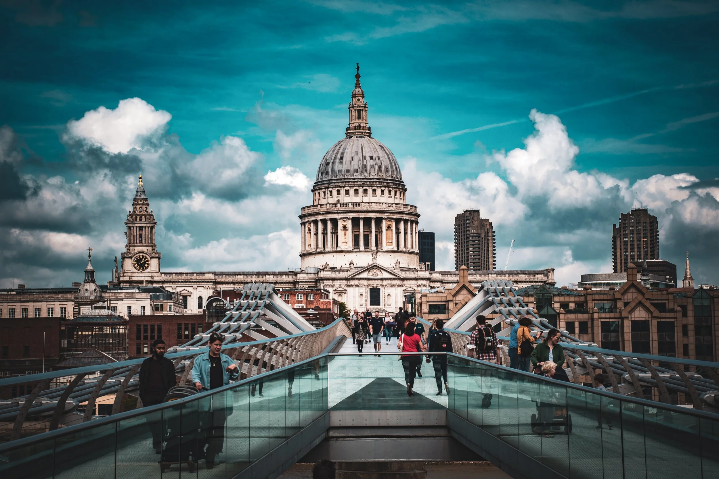 People walking on Millennium Bridge with St. Paul's Cathedral in London in the background under a partly cloudy sky.