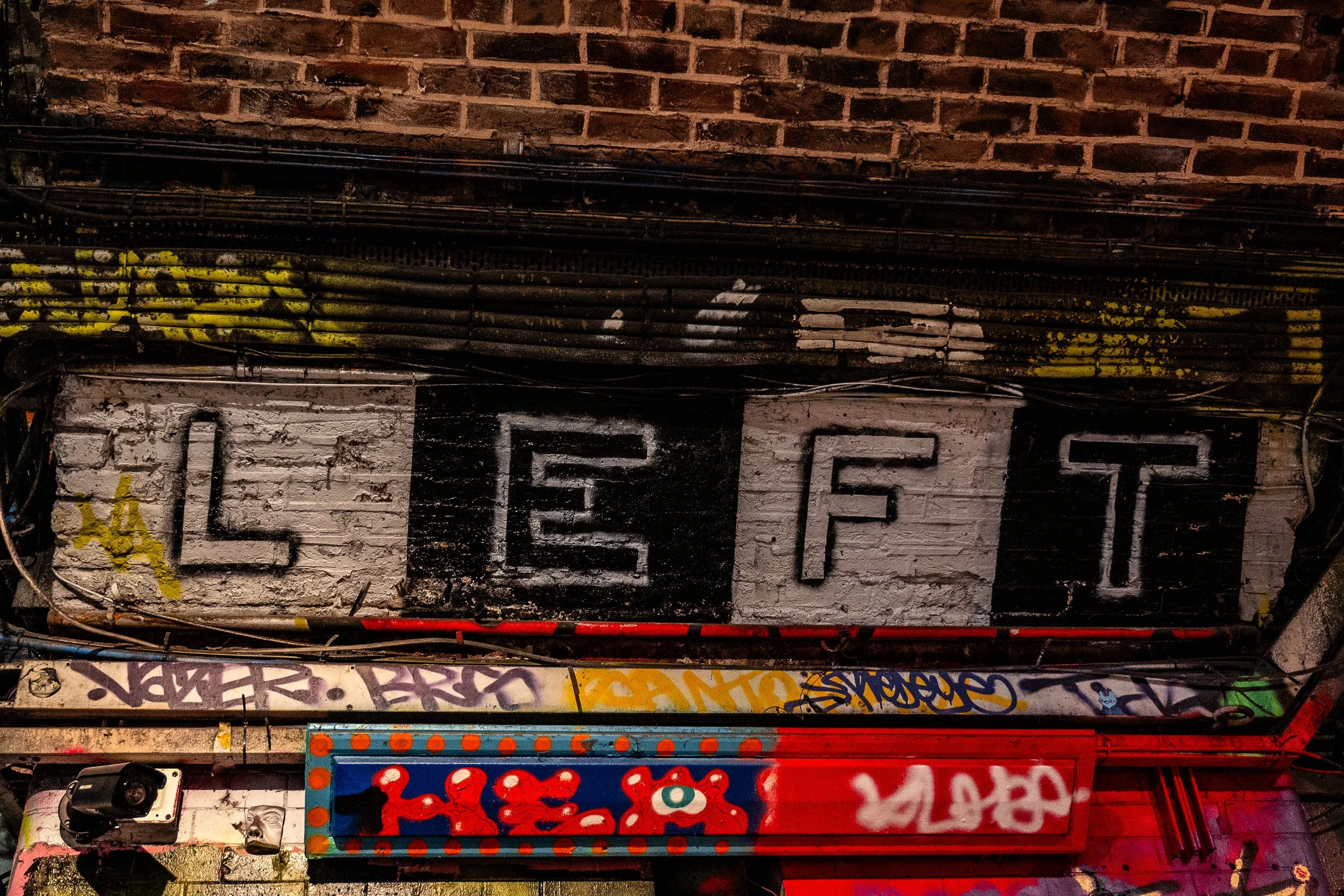 A brick wall with graffiti and a painted sign that spells out 'LET' in large letters. The sign has a black and white background, with the letters painted in a stencil style.
