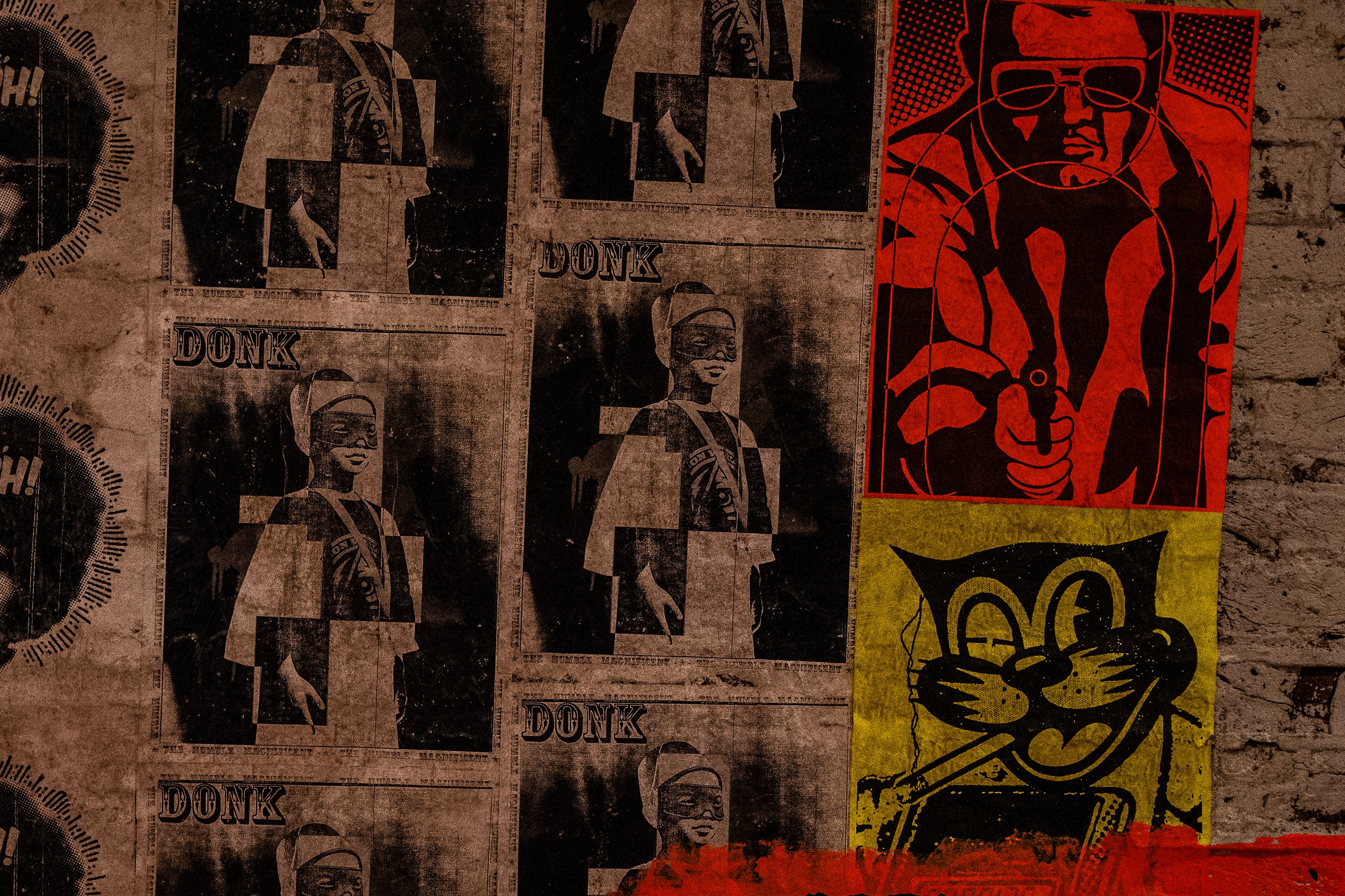 Street art posters on a brick wall, including black and white posters with a woman wearing glasses and the word 'DONK', a red poster with a stylized face, and a yellow poster with a cartoon cat.