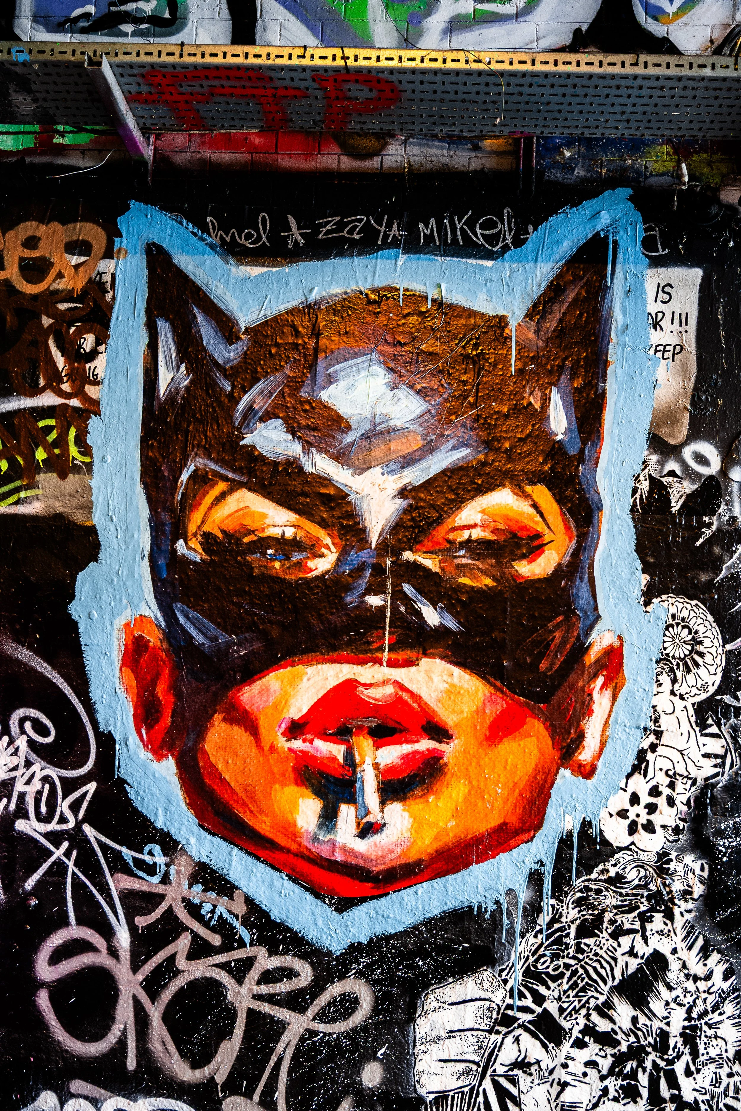 Graffiti street art of a woman's face wearing a Batman mask, with bright red lips, painted on a wall with various tags and doodles around it.