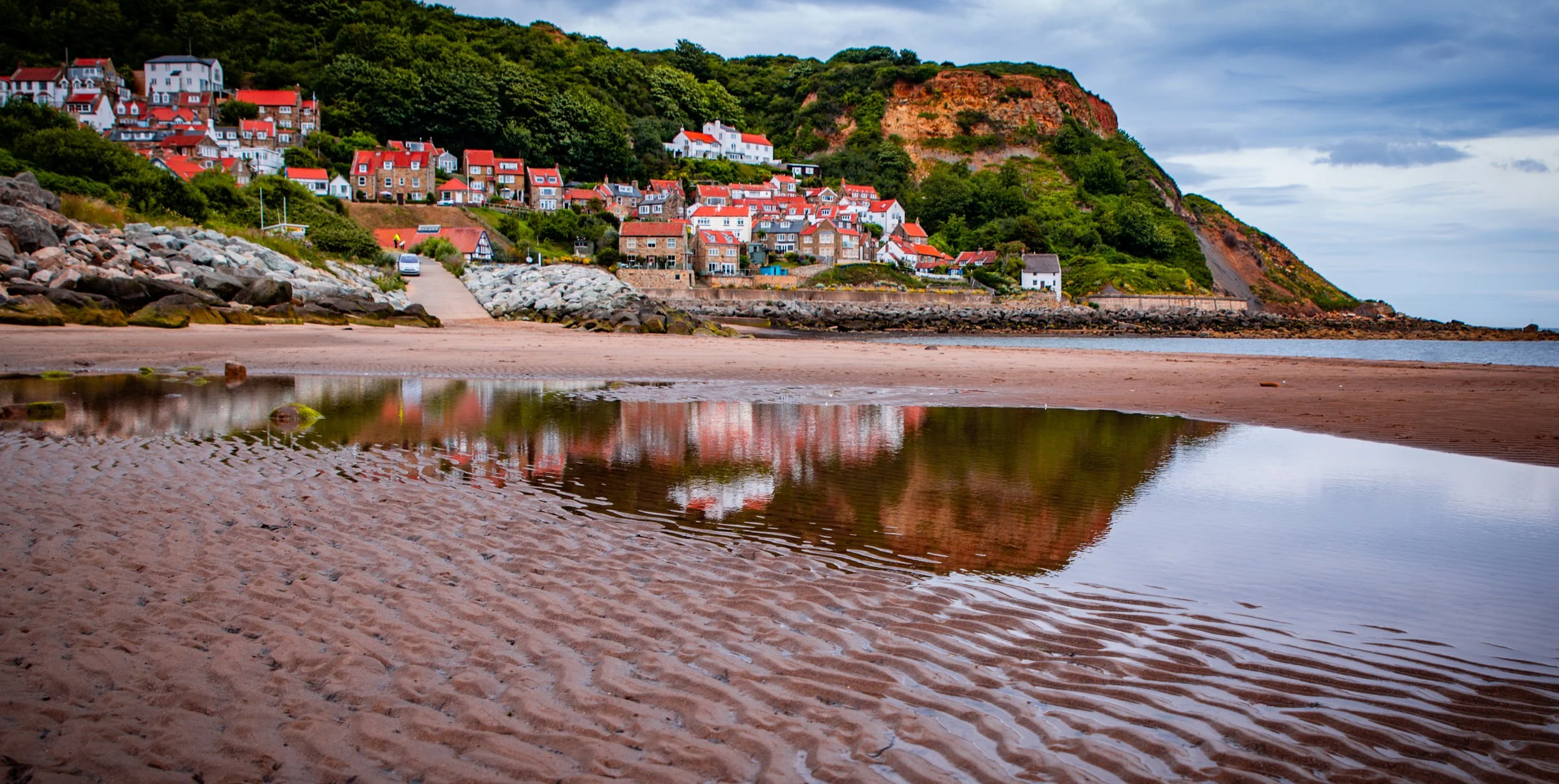 A scenic coastal village with colorful houses on a hillside, visible from a sandy beach with rippled sand and a small tide pool reflecting the houses and sky.