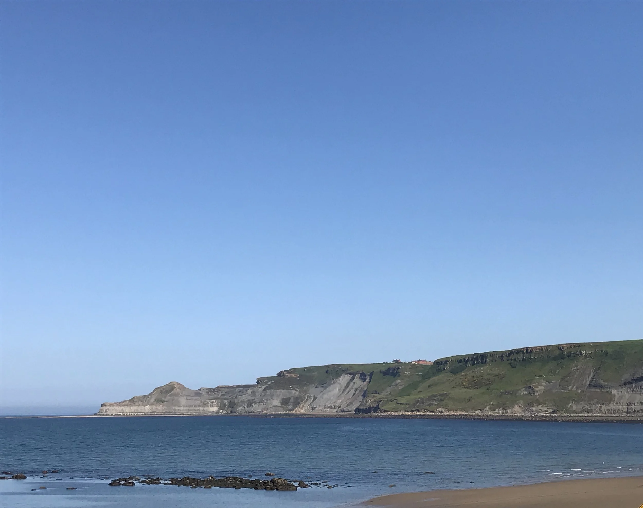 Coastal landscape with green cliffs, sandy beach, and blue sky over calm ocean water.