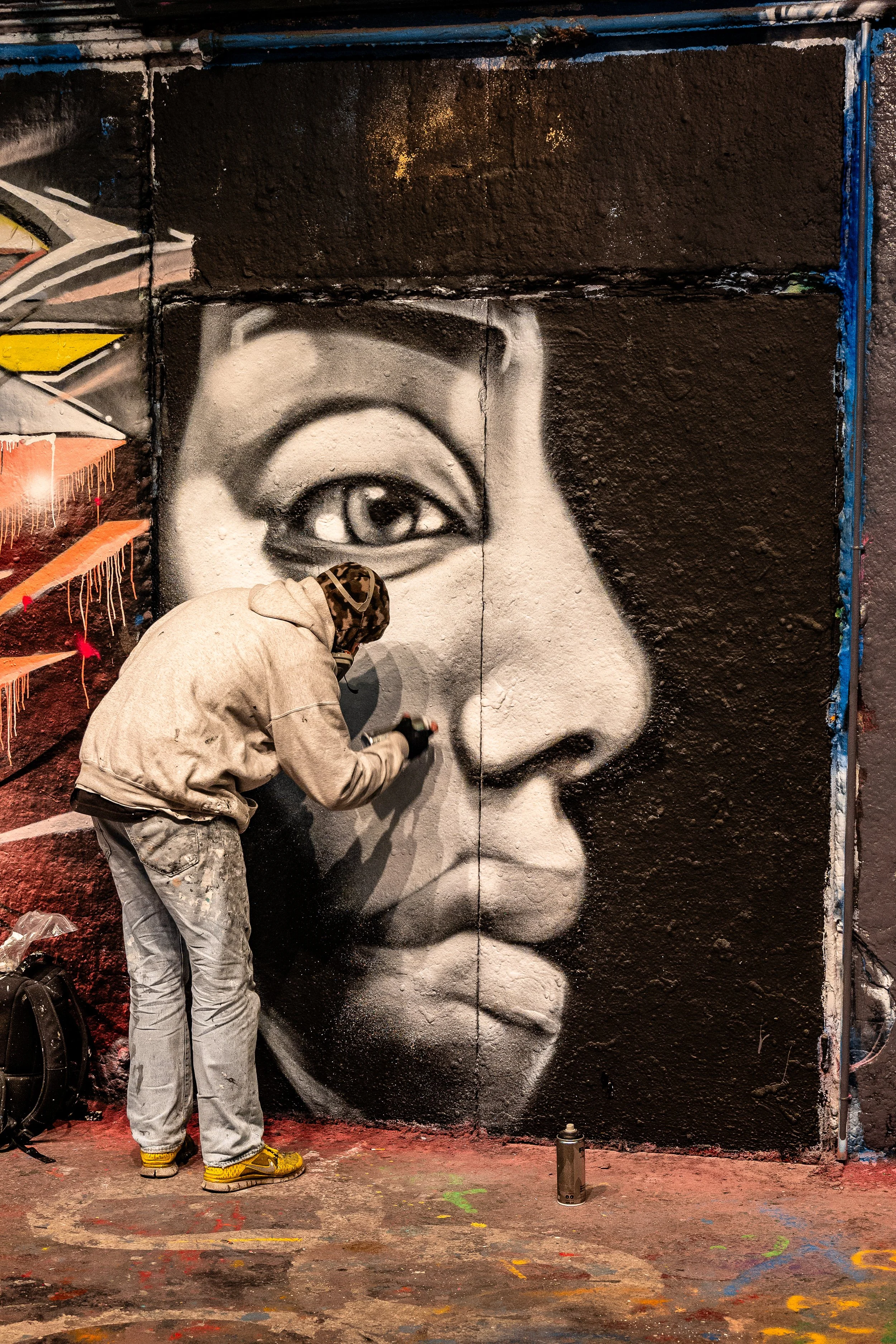 An artist in a hoodie and jeans creating a large black-and-white portrait of a woman's face on a graffiti-covered wall.