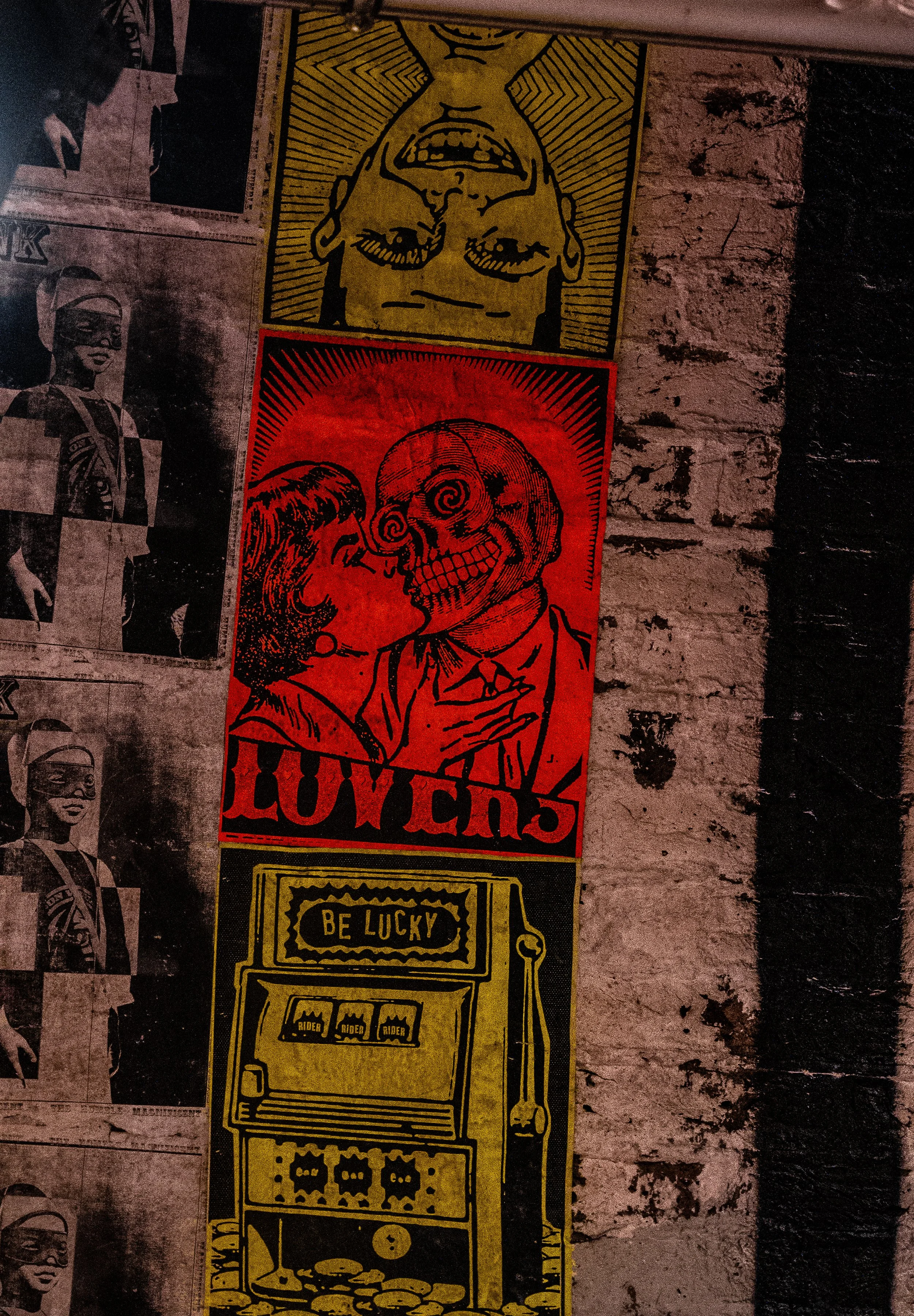 Colorful street art posters on a brick wall. The top poster features an upside-down face with a yellow background. The middle poster shows a person kissing a skeleton, with the word "LOVERS" at the bottom, on a red background. The bottom poster illus