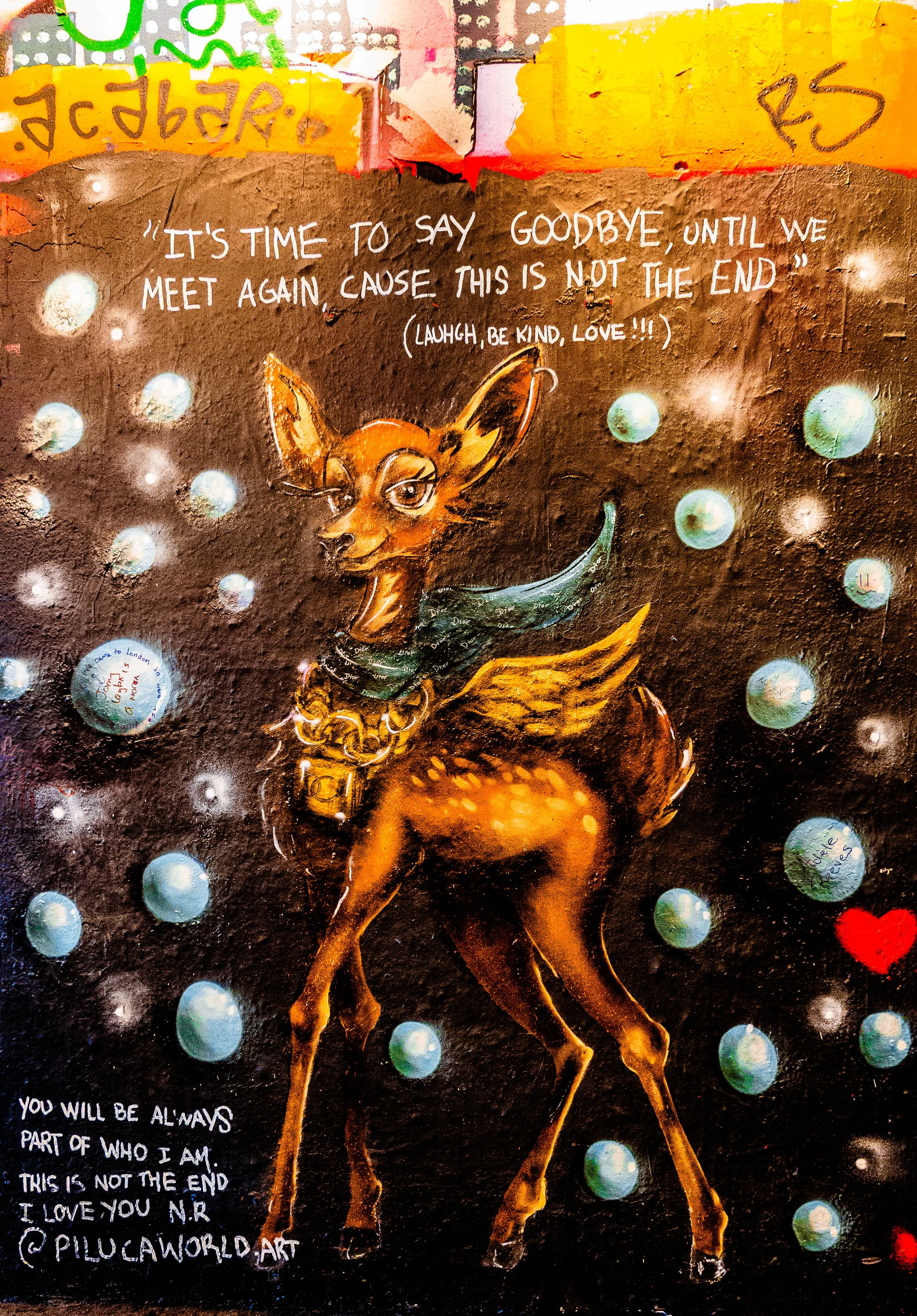 Colorful graffiti art featuring a stylized, cartoon-like deer with an orange and brown body, large eyes, and a green scarf around its neck. The background is dark with glowing orbs and a red heart. The artwork includes a quote about saying goodbye an