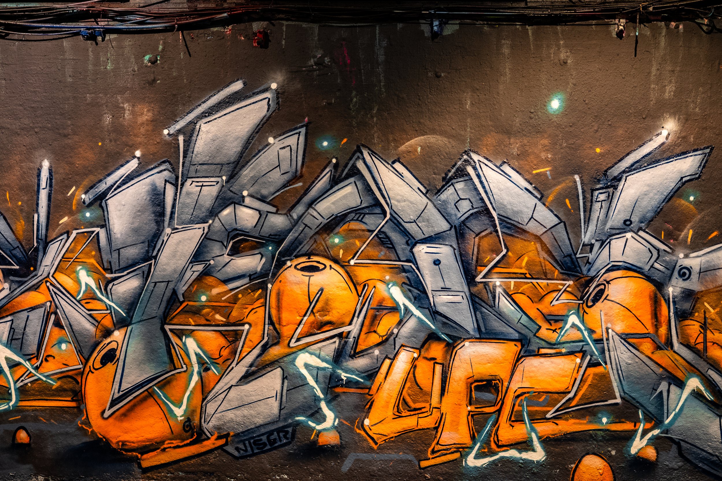 Colorful graffiti art on a wall featuring abstract shapes in black, gray, orange, and white with teal accents and glowing effects.