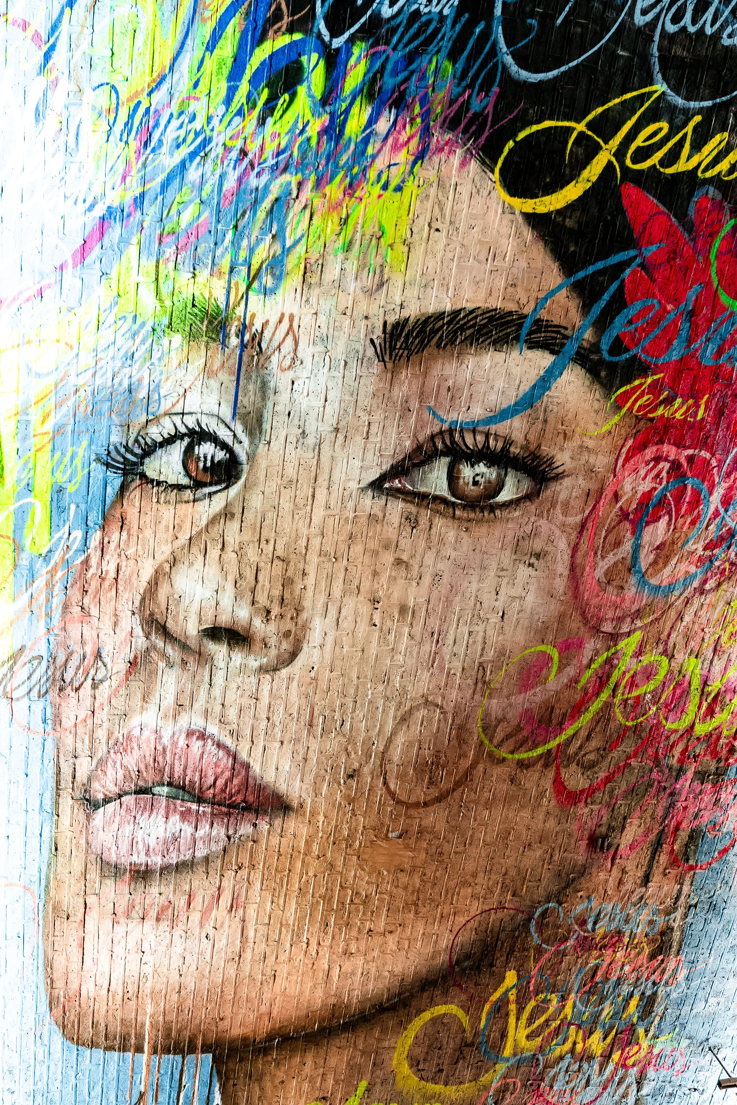 A mural of a woman's face with brown eyes, dark eyebrows, and pink lips, overlaid with colorful, cursive graffiti writing.