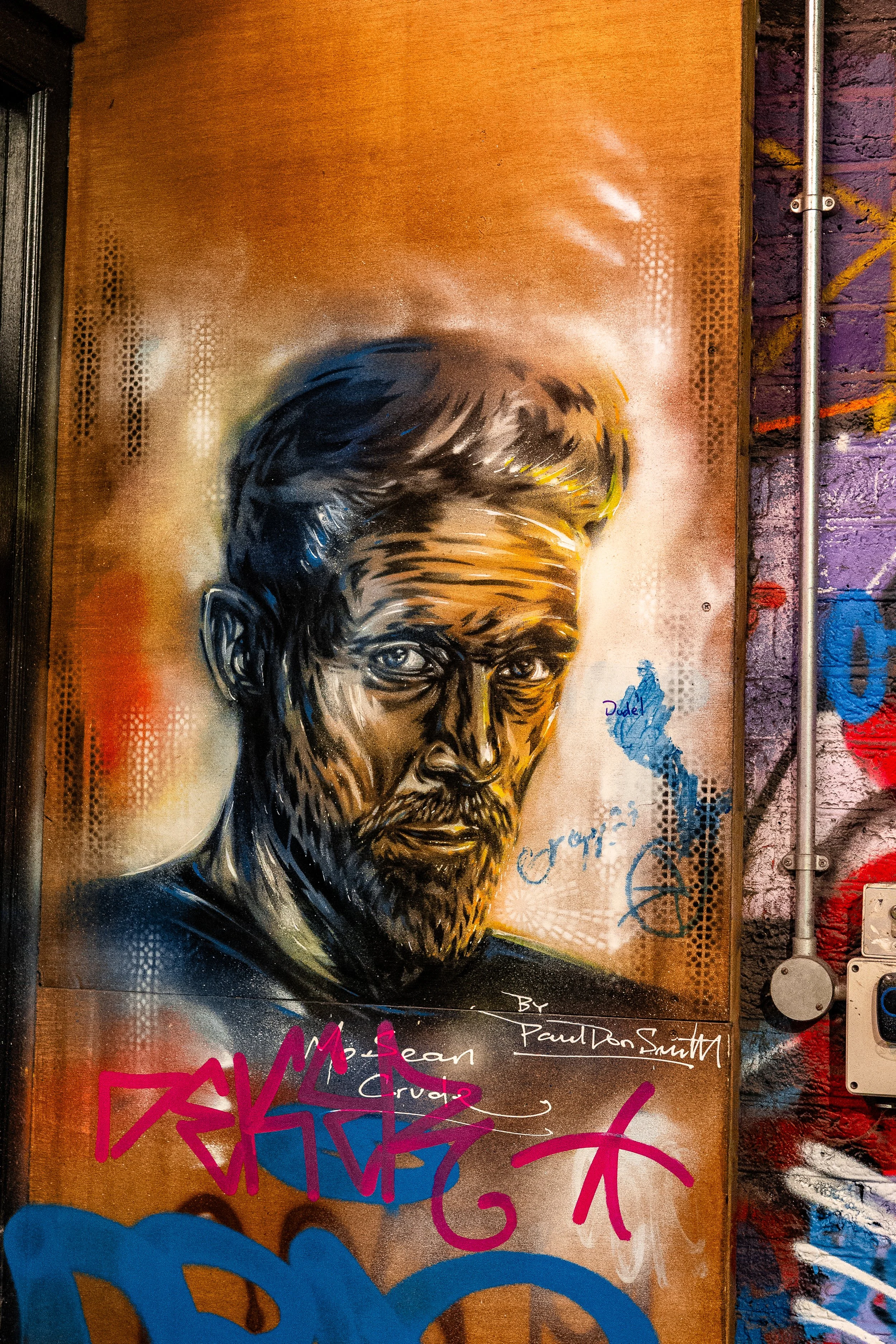 Graffiti mural of a man's portrait with short hair and beard, painted on a wall with orange and purple background, including signatures and tags.