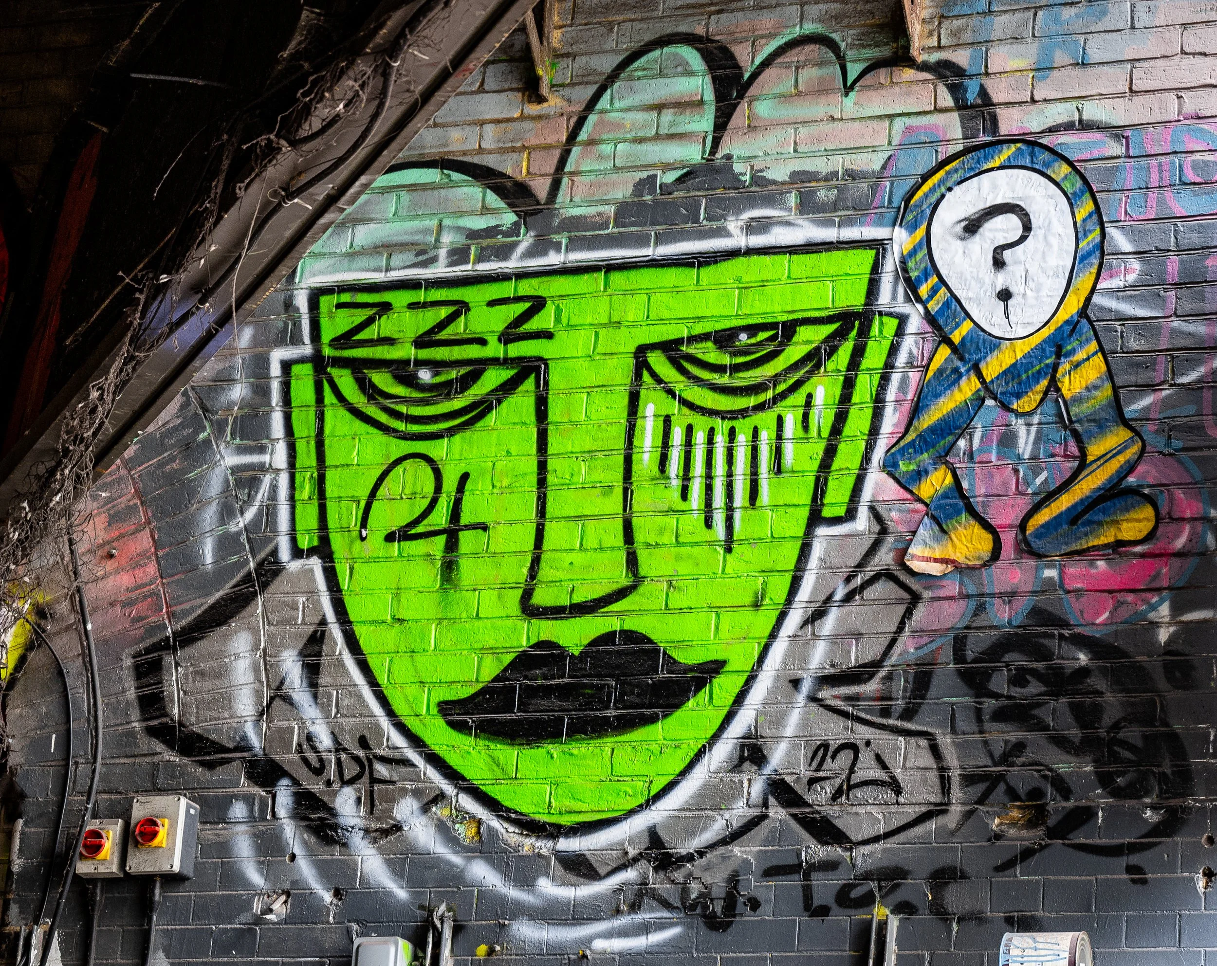 Graffiti art depicting a green face with tired eyes and black lips, with a small hunched figure with a white speech bubble containing a question mark. The background is a brick wall with various colors and tags, and there are electrical outlets and w